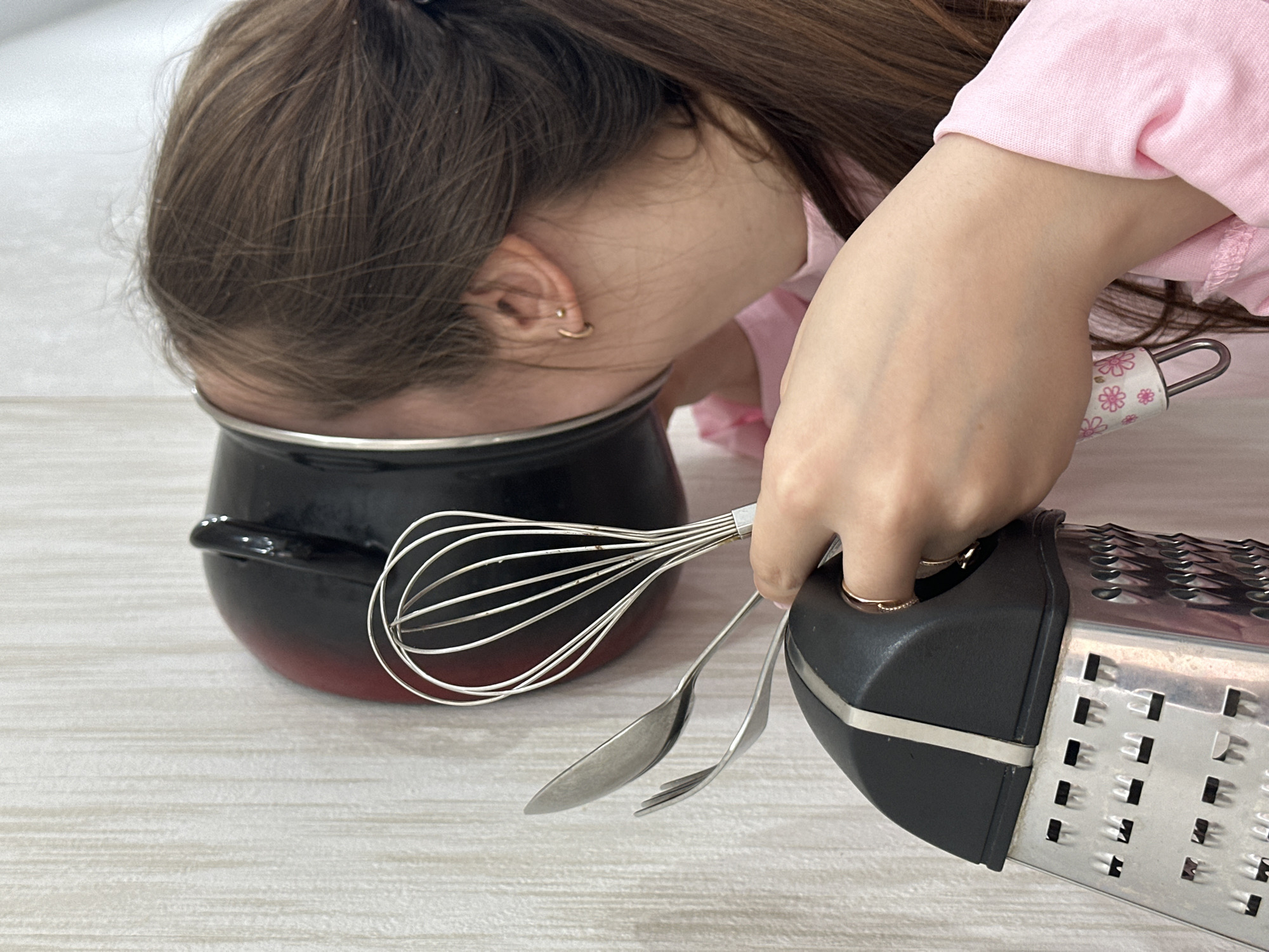 A child's head is stuck in a red and black pot on a light-colored surface. To the right of the pot, a hand holds a grater along with a whisk, spoon, and fork. The location is Chaikovsky, Russia.
