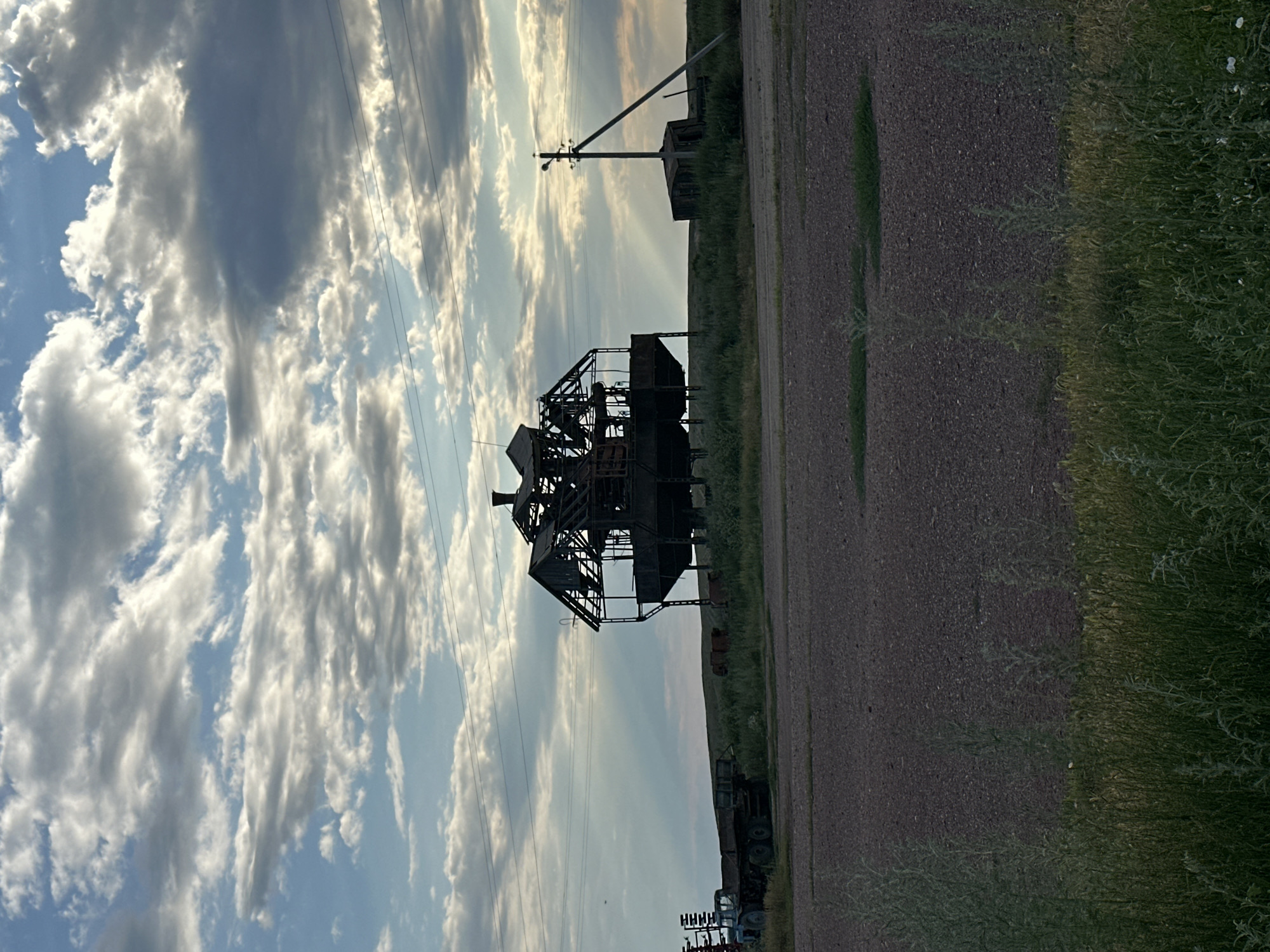 The image depicts an abandoned industrial structure, possibly a former processing plant or factory, set against a dramatic sky filled with dramatic clouds illuminated by rays of sunlight. The structure is skeletal and in disrepair, made of metal and wood, with a distinctive multi-tiered design. In the foreground, there's a flat, gravelly area. To the left, a blue tractor trailer is parked. The background features a grassy field with a small, dilapidated shed and a utility pole. The overall impression is one of rural decay and desolation. The specific location in Bayagiz, Kazakhstan, is not visually identifiable from the image itself, but it is presented as the setting.