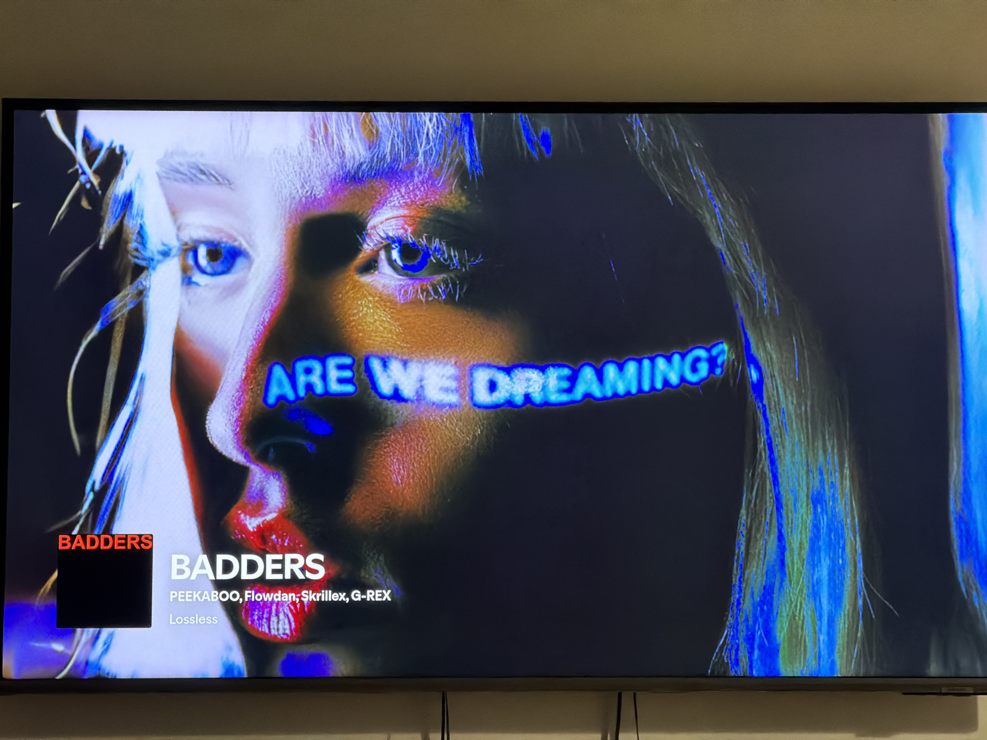 The image shows a close-up of a television screen displaying a music video. The screen features a stylized portrait of a person's face with prominent blue and red lighting effects. Overlayed on the image are text elements. In the bottom left corner, a red rectangle contains the word "BADDERS" in white text, with a black square below it. To the right of this, the word "BADDERS" appears again in larger white text, followed by "PEEKABOO, Flowdan, Skrillex, G-REX" and "Lossless" in smaller white text. Above the face, the text "ARE WE DREAMING?" is visible in glowing blue letters. The background of the screen is dark. The television is mounted on a wall, and the top edge of the wall is visible above the screen. The image is taken from an angled perspective, with some glare or reflections on the screen. There is no discernible information in the image that would indicate its location in Sosnowiec, Poland.
