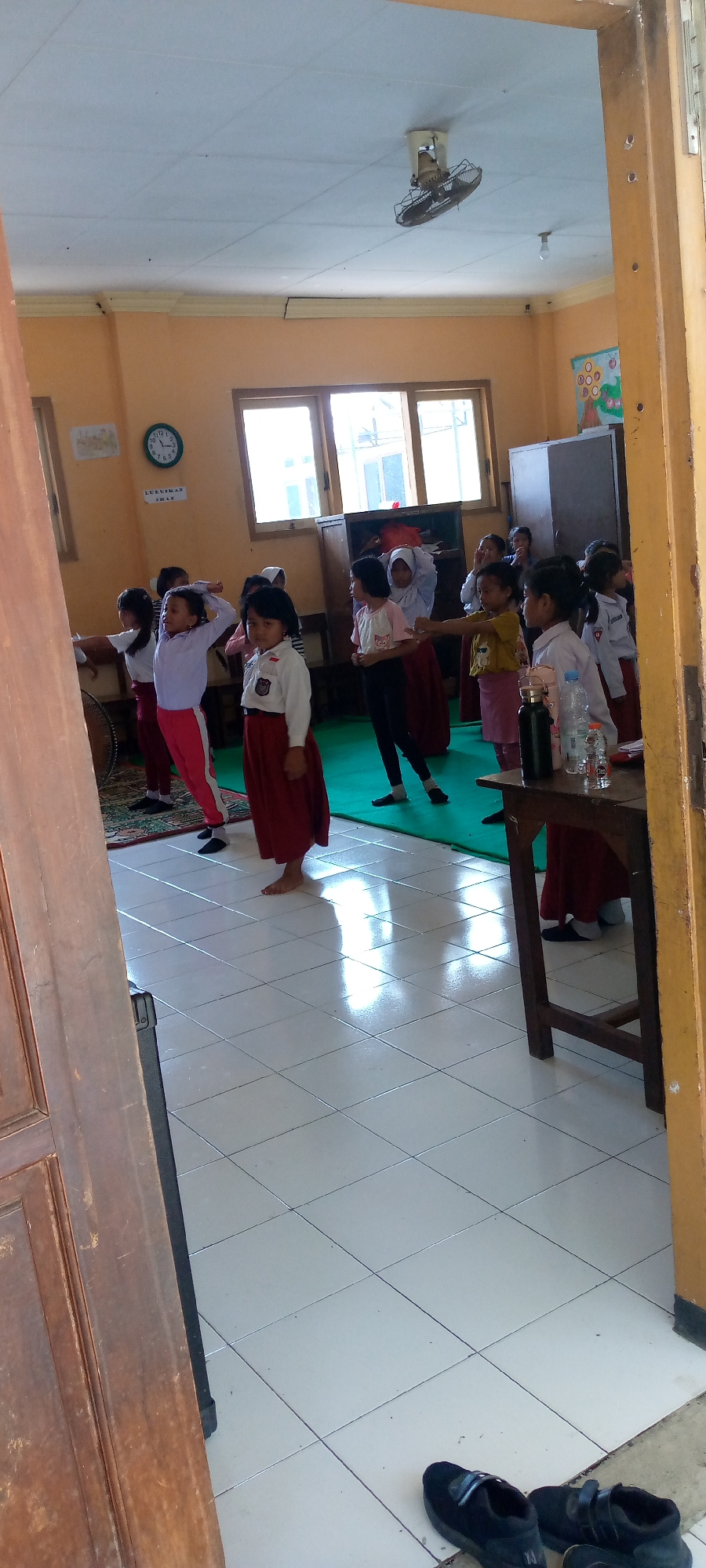 In Pacing, Indonesia, a group of young female students, dressed in school uniforms consisting of white shirts and red or maroon skirts/pants, are gathered in a classroom for what appears to be a physical education activity or dance lesson. The room has light orange walls and large windows that allow natural light to enter, illuminating the tiled floor and a green mat on the floor. A ceiling fan is visible, suggesting the room might get warm. On one wall, a round analog clock is hung, showing a time around 2:10. Various children are positioned in different stances, some with arms raised, indicating movement or instruction. A wooden table is present in the foreground with several water bottles and other items on it. The overall scene suggests an active and educational environment for young learners in Indonesia.