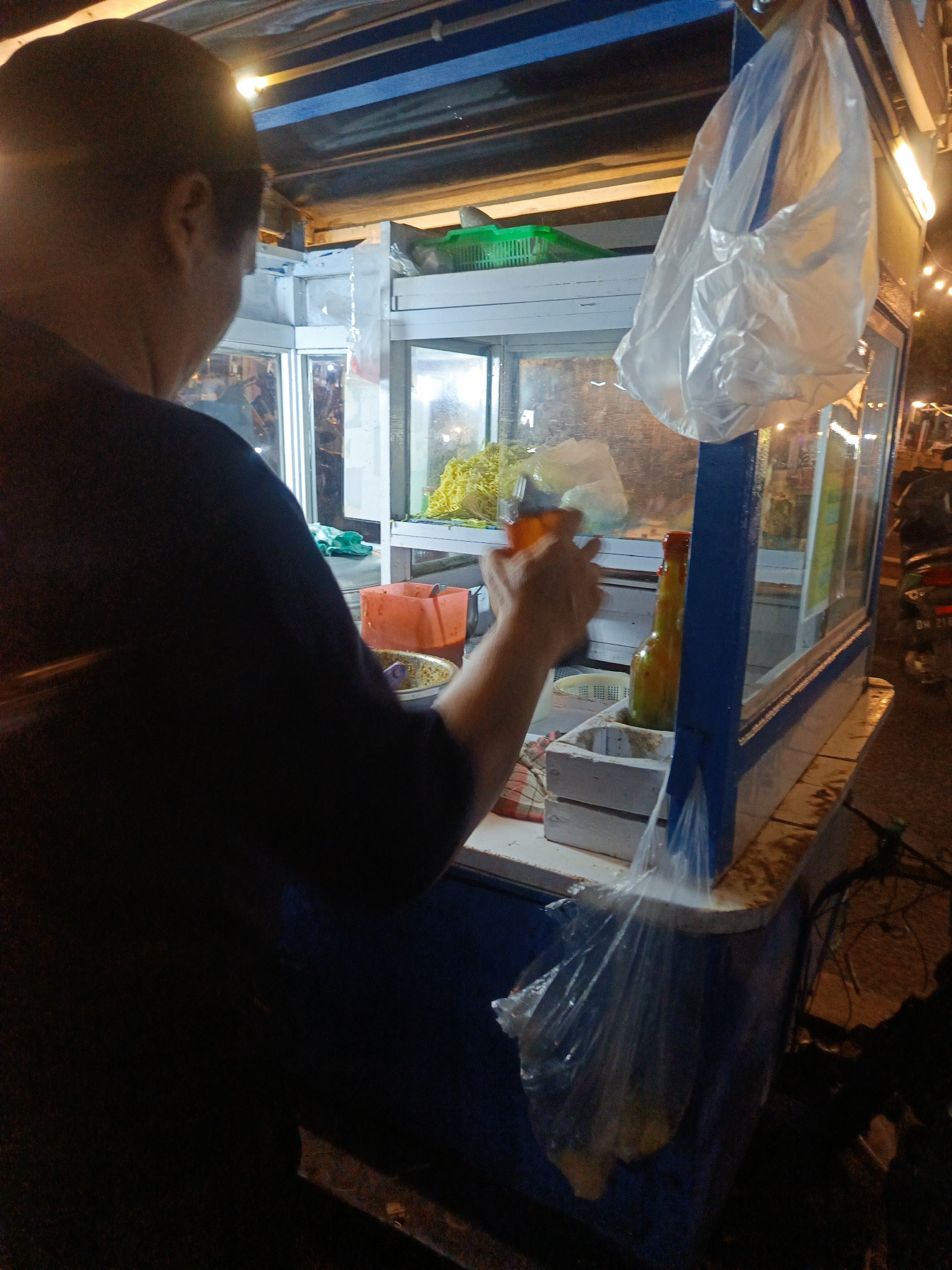 A person is seen from behind and to the left, wearing a dark shirt, standing at a food stall in Luwoo, Indonesia. The food stall appears to be a mobile cart with a glass display case. Inside the display, there are yellow noodles and other food items. An orange container and a bottle with liquid are visible on the counter. A plastic bag hangs from the top of the stall. The scene is lit by artificial lights, suggesting it is evening or nighttime. A motorcycle is parked to the right of the stall.