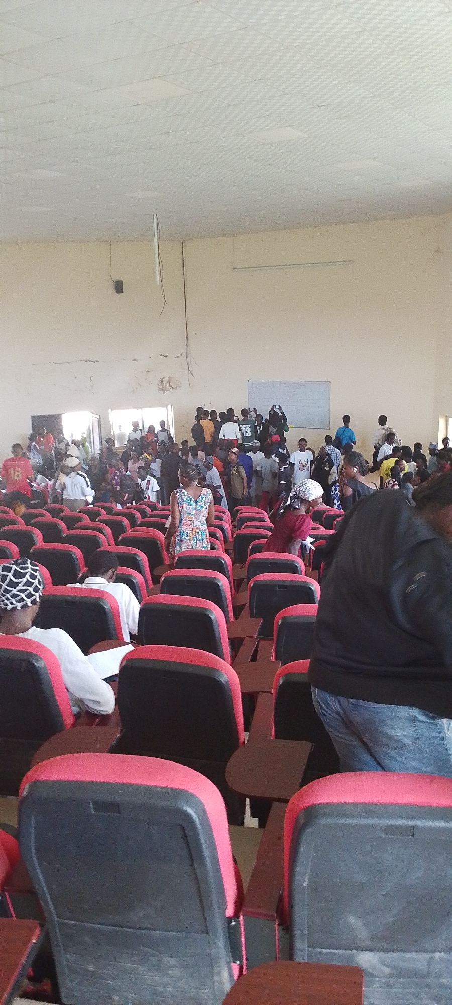 The image depicts an indoor scene, likely a lecture hall or auditorium, with rows of red and black seating. A group of people are gathered in the center of the room, some standing and interacting, while others are seated or in the process of sitting down. The ceiling is white and appears to be made of acoustic tiles, with some visible wires or cables hanging down. The walls are a light, neutral color and show signs of wear. There is a whiteboard on the back wall. The presence of numerous people suggests an event or class is taking place. The reference to Gumel, Nigeria, places this scene within that geographical context.