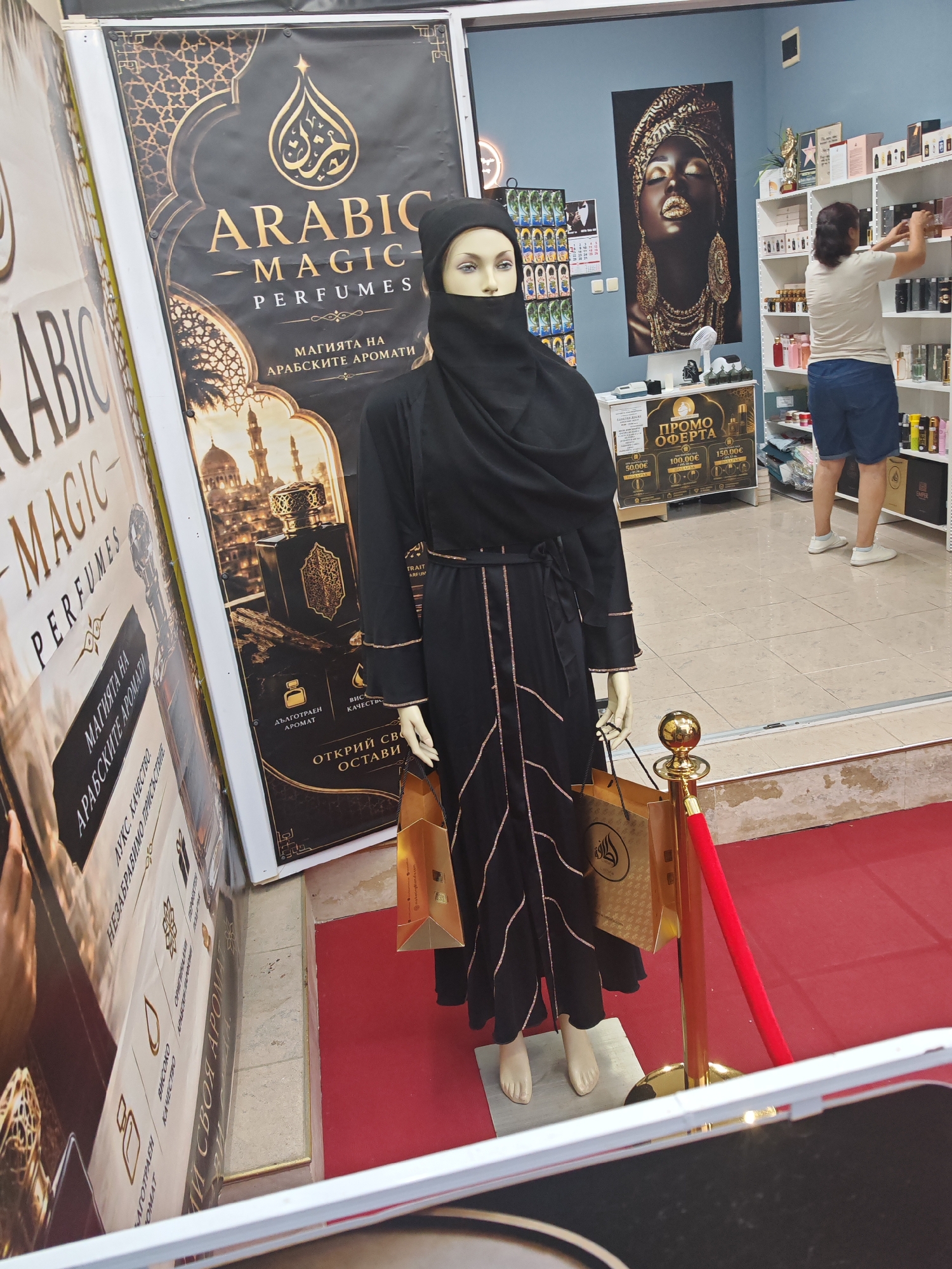 The image shows the interior of a perfume shop located in Kiten, Bulgaria. A mannequin dressed in traditional Islamic attire is displayed prominently in the foreground. The mannequin is wearing a black hijab and abaya, and is holding two golden shopping bags. Behind the mannequin, there is a large poster advertising "Arabic Magic Perfumes," featuring ornate Arabic calligraphy and images of mosques and perfume bottles. To the right of the mannequin, a woman is browsing shelves stocked with various perfume bottles. The shop has a red carpet and golden stanchions with red ropes, creating a sense of luxury. The walls are decorated with posters and shelves displaying merchandise. The overall ambiance suggests a retail environment focused on oriental-themed perfumes.