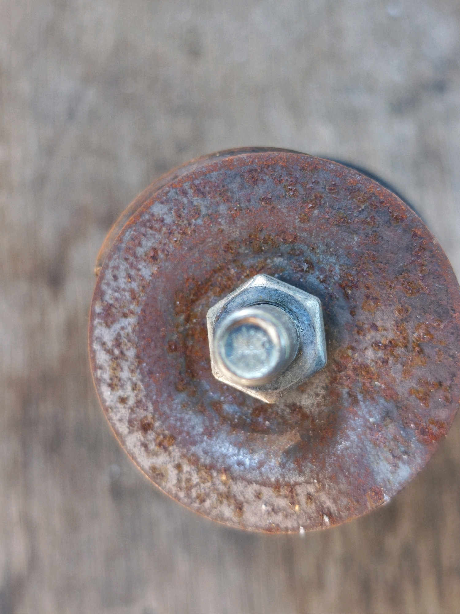 The image is a close-up, top-down view of a rusty, circular metal object secured by a bolt and nut. The object appears to be part of a larger mechanism, possibly industrial or mechanical equipment. The metal is heavily oxidized, displaying shades of brown, orange, and blue-grey. The bolt and nut in the center are metallic and appear to be in better condition than the surrounding rusted metal. The background is a blurred, light brown, textured surface, likely wood or concrete, providing a neutral backdrop that emphasizes the subject. No people or specific environmental context beyond the immediate objects are visible. The location is not determinable from the image itself; it is a generic depiction of a weathered metal component.