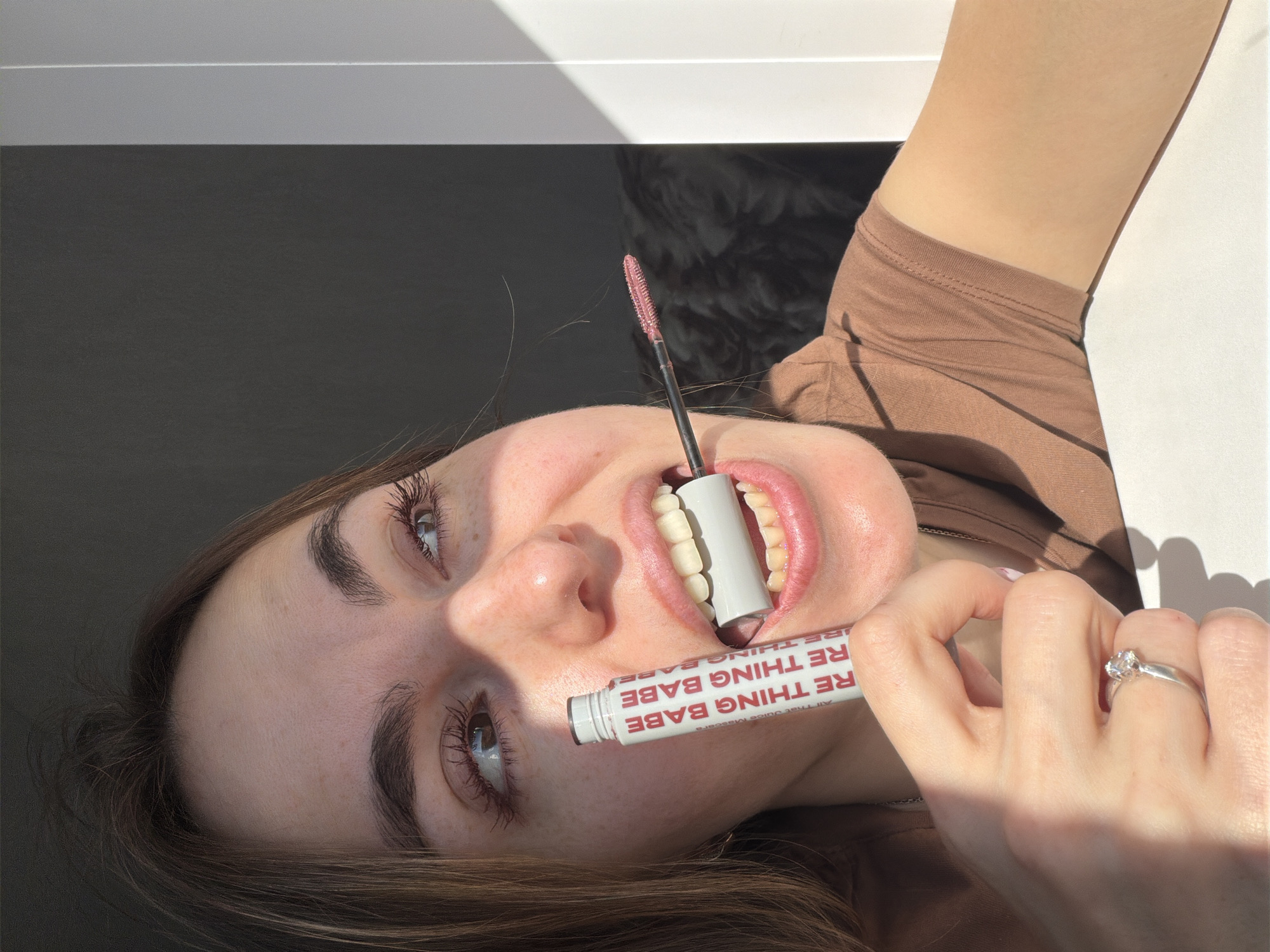 A woman is posing with a tube of mascara. The tube, labeled "RE THING BABE All That Juice Mascara," is held in her mouth and in her left hand, which has a diamond ring on the ring finger. The brush from the tube is out and upright. She is wearing a brown shirt. The location and city of Sarapul, Russia cannot be determined from the image.