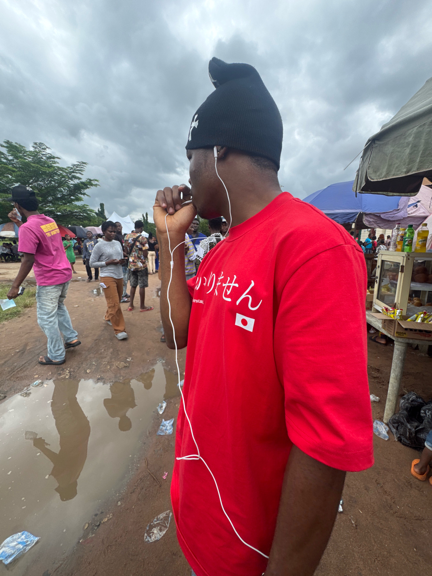 The image depicts an outdoor scene, likely a marketplace or gathering, under an overcast sky. The foreground features a person from the chest down wearing a bright red t-shirt with white Japanese characters and a Japanese flag emblem. White earphone wires are visible, one tucked behind the ear and the other dangling down the front of the shirt. The ground is uneven and muddy, with several large puddles reflecting the sky and surrounding figures.
In the mid-ground, various individuals are present. To the left, a person in a pink t-shirt and light-colored jeans walks away from the camera, holding what appears to be a white piece of paper. Further back, a woman in a grey long-sleeved shirt and brown pants looks at her phone. Other people are scattered throughout the scene, some appearing to be vendors under tarps or umbrellas. The background shows more people, tents, and a stall with various items, including bottled drinks and boxed goods. The overall atmosphere suggests a busy, perhaps somewhat disorganized, public event in a rural or semi-urban setting, consistent with a location like Ihiagwa, Nigeria.