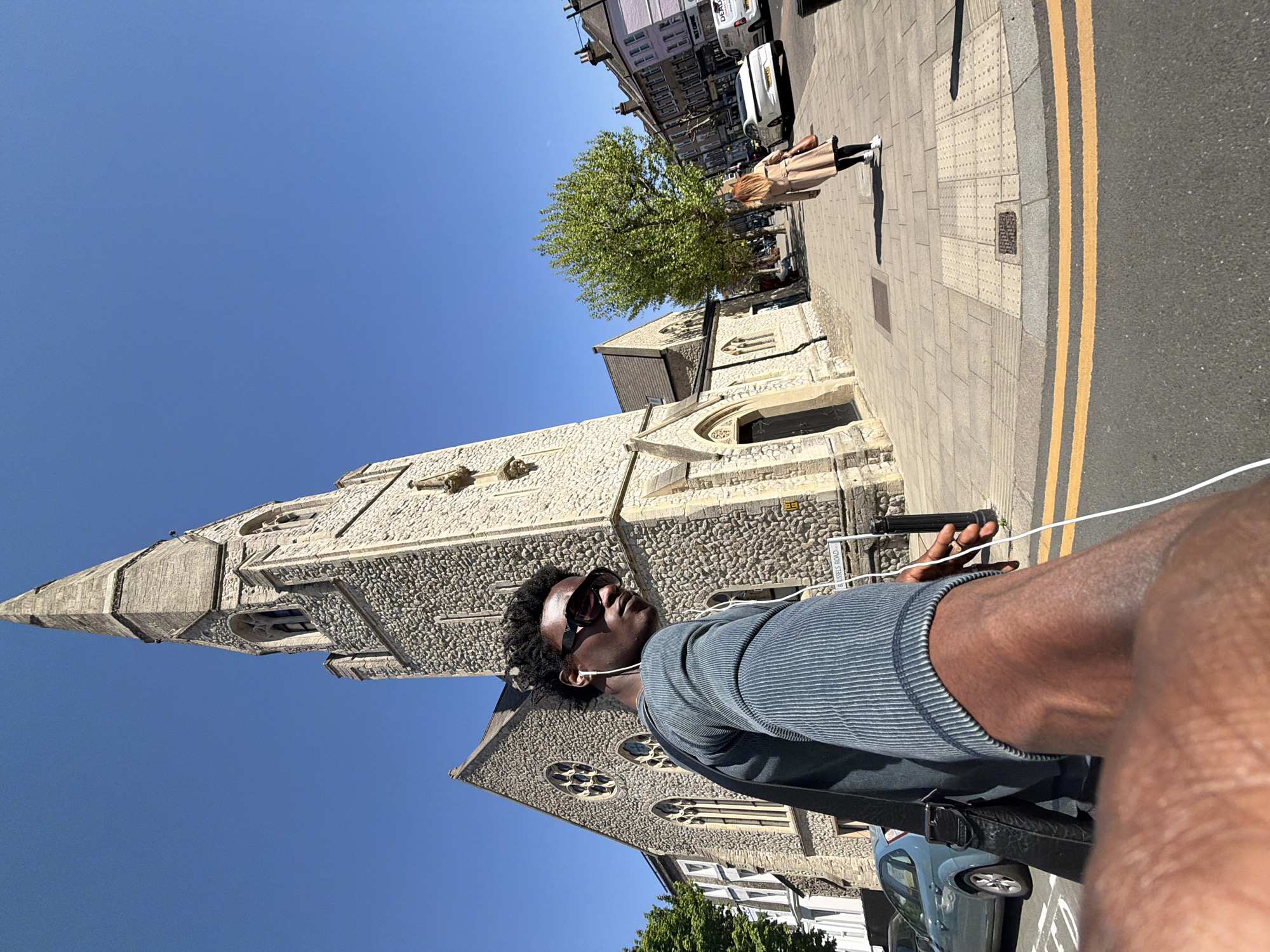 A photograph taken on a sunny day from a low angle shows a Black man wearing sunglasses and headphones, standing on a city street in London, United Kingdom. The man is wearing a dark blue horizontally-striped shirt and a black shoulder bag. A tall church made of light grey stone with a pointed steeple is behind him. The street has a grey sidewalk and a black street with double yellow lines. In the background, there are other buildings, trees, and pedestrians. A street sign reads "RUSSELLS ROAD". A parked blue car is visible.