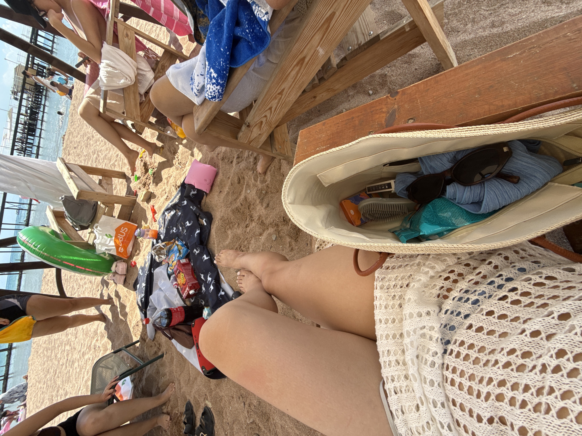 The image depicts a beach scene in Sary-Oy, Kyrgyzstan, on a sunny day. In the foreground, a person's legs are visible, stretched out on the sand, covered by a white, crocheted garment. A beige tote bag sits next to the legs, containing a blue scarf, sunglasses, and a hairbrush. In the middle ground, various beach items are scattered on the sand, including a red Coca-Cola bottle, a patterned cloth, a pink inflatable pillow, and a white plastic bag with the "Ebus" logo. Two green and yellow inflatable rings are also present, along with a pair of pink child-sized shoes. In the background, several people are seated on beach chairs, and others are visible in the shallow water near a pier. The environment is sandy, with clear blue water and a bright sky.