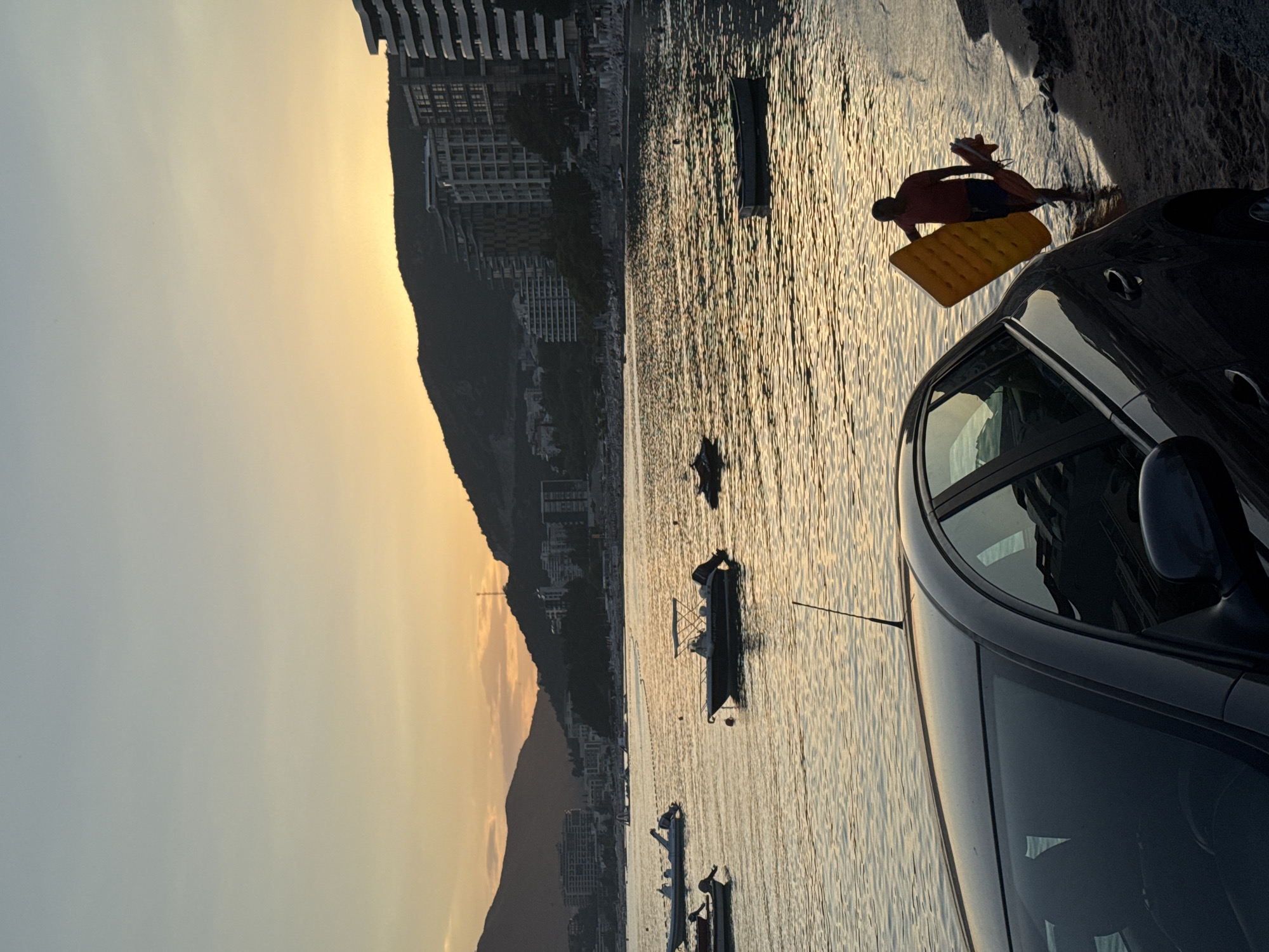 A man is walking into the water on a beach in Rafailovići, Montenegro, carrying a yellow inflatable mattress and a red flotation device. The sun is setting, casting a warm glow over the scene. Several boats are moored in the calm sea. In the foreground, the rear of a dark-colored car is visible, parked close to the water's edge. The coastline is lined with buildings and hills.