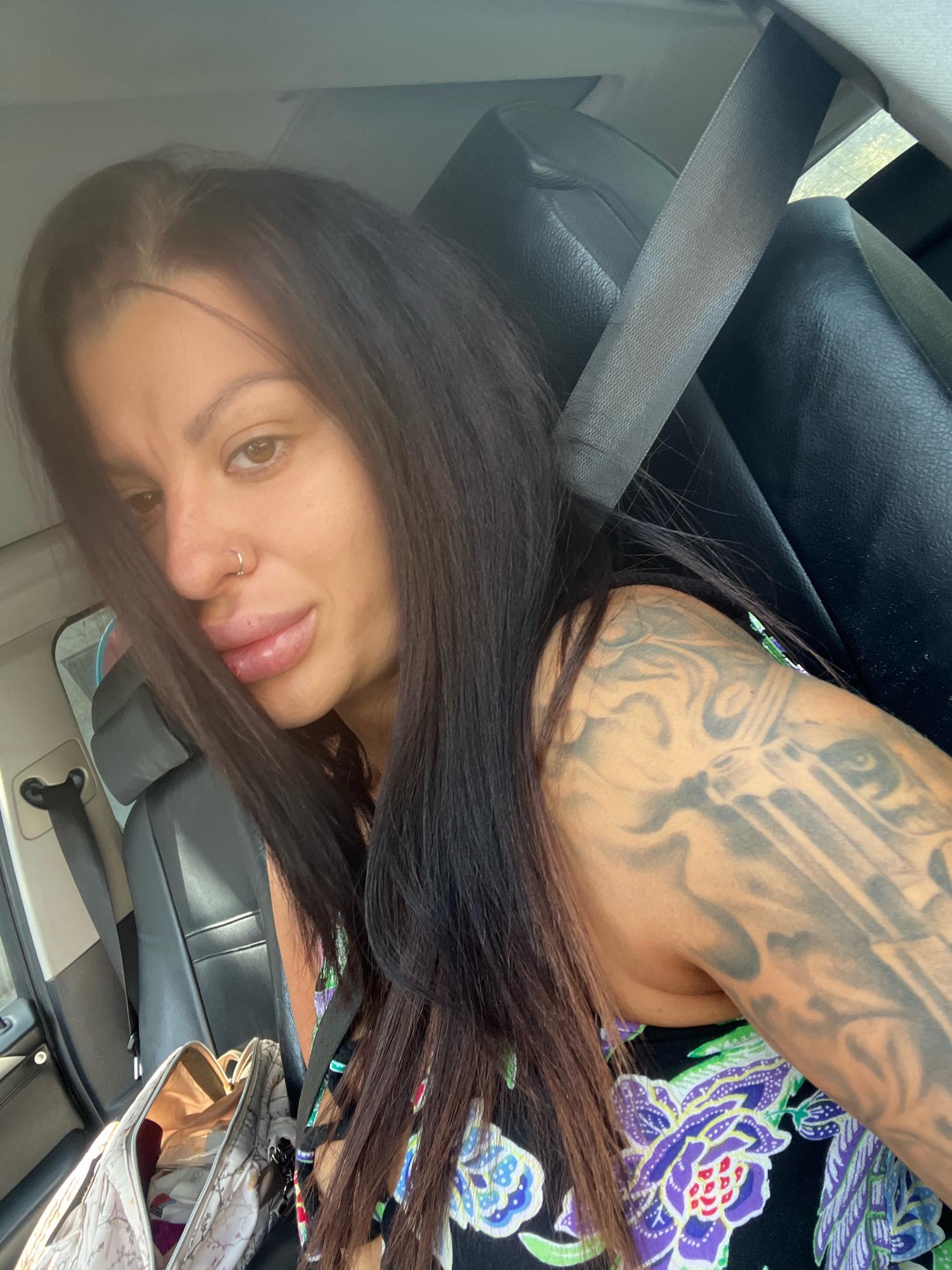A woman with long, dark hair is sitting in the passenger seat of a car, her body turned towards the camera. She is wearing a black dress with a colorful floral pattern. Her left arm is visible, covered in a large tattoo of a gun and swirling smoke. A seatbelt is across her chest. In the foreground, to the left, a beige and white patterned duffel bag is open, with items inside. The background shows the car's black leather interior and a glimpse of the outdoors through a window. The image is an indoor shot, taken within a vehicle. There is no information in the image to suggest the location is Plovdiv, Bulgaria.