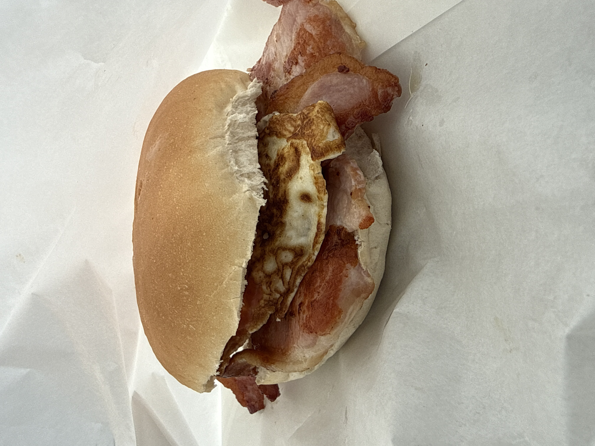 A close-up, eye-level shot shows a bacon and egg sandwich. The sandwich is presented in a white paper wrapper, with some of the paper folded up around it. The sandwich itself is made with a soft, golden-brown bun, split horizontally. Inside, two slices of fried egg, with a slightly crispy edge, are visible. These are layered with several strips of cooked bacon, which have a streaky, reddish-pink hue and some crispy edges. The bacon extends slightly beyond the bun on both sides. The background consists of crinkled white paper, suggesting the sandwich is being held or presented in disposable packaging. The lighting is bright, creating soft shadows and highlighting the textures of the food and paper. The setting is a street food stall or cafe in Tewkesbury, United Kingdom, likely on a street such as High Street or Church Street.