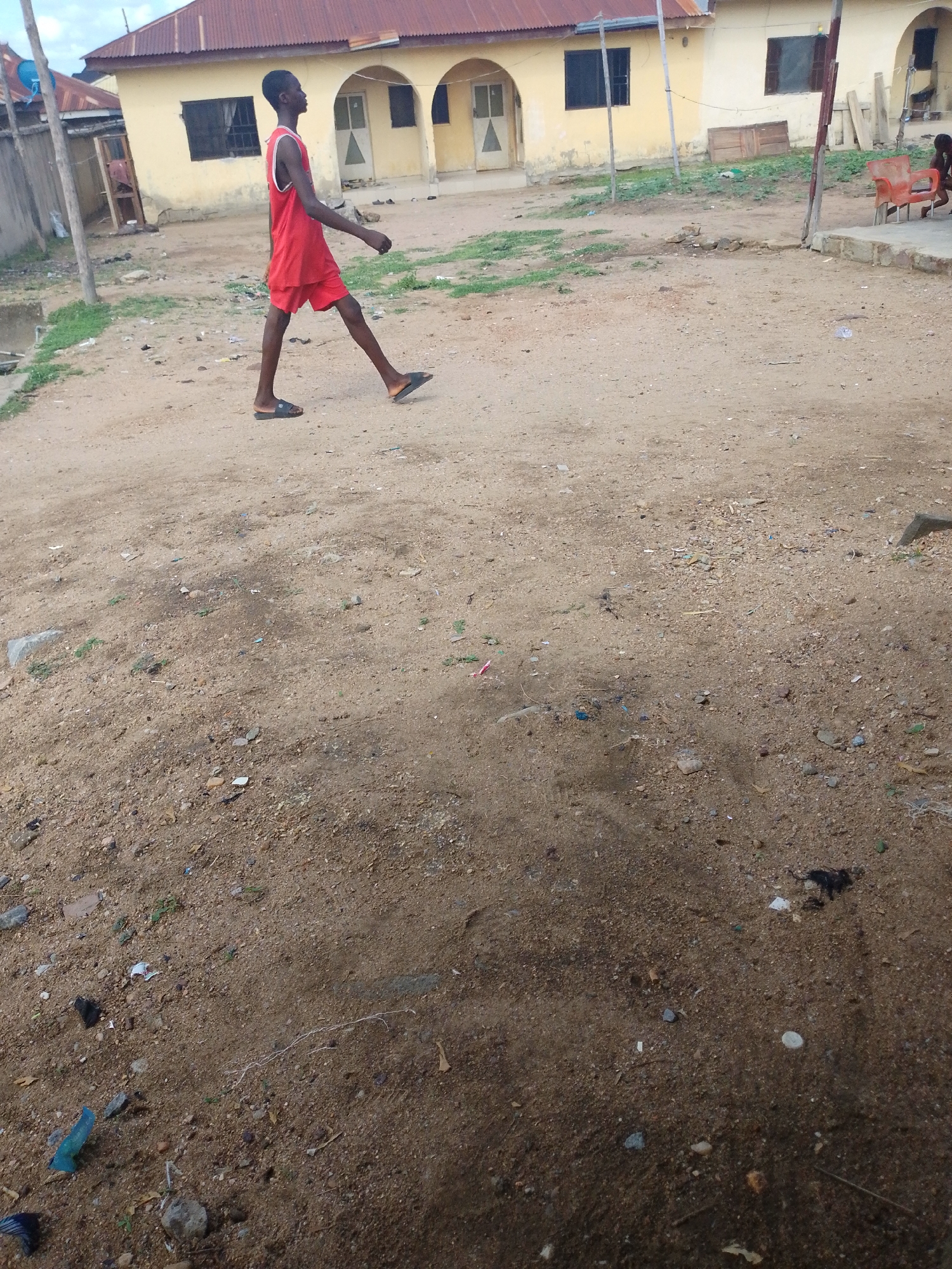 A young man in a red athletic outfit walks from left to right across a dirt-covered area in Lokoja, Nigeria. The location appears to be a village area. In the background are buildings, mostly one-story, with red or yellow painted walls and corrugated metal roofs. Some of the buildings have arched doorways. A wooden pole stands to the left. Debris litters the ground, including small rocks, leaves, and bits of plastic or other refuse. The sky is overcast.