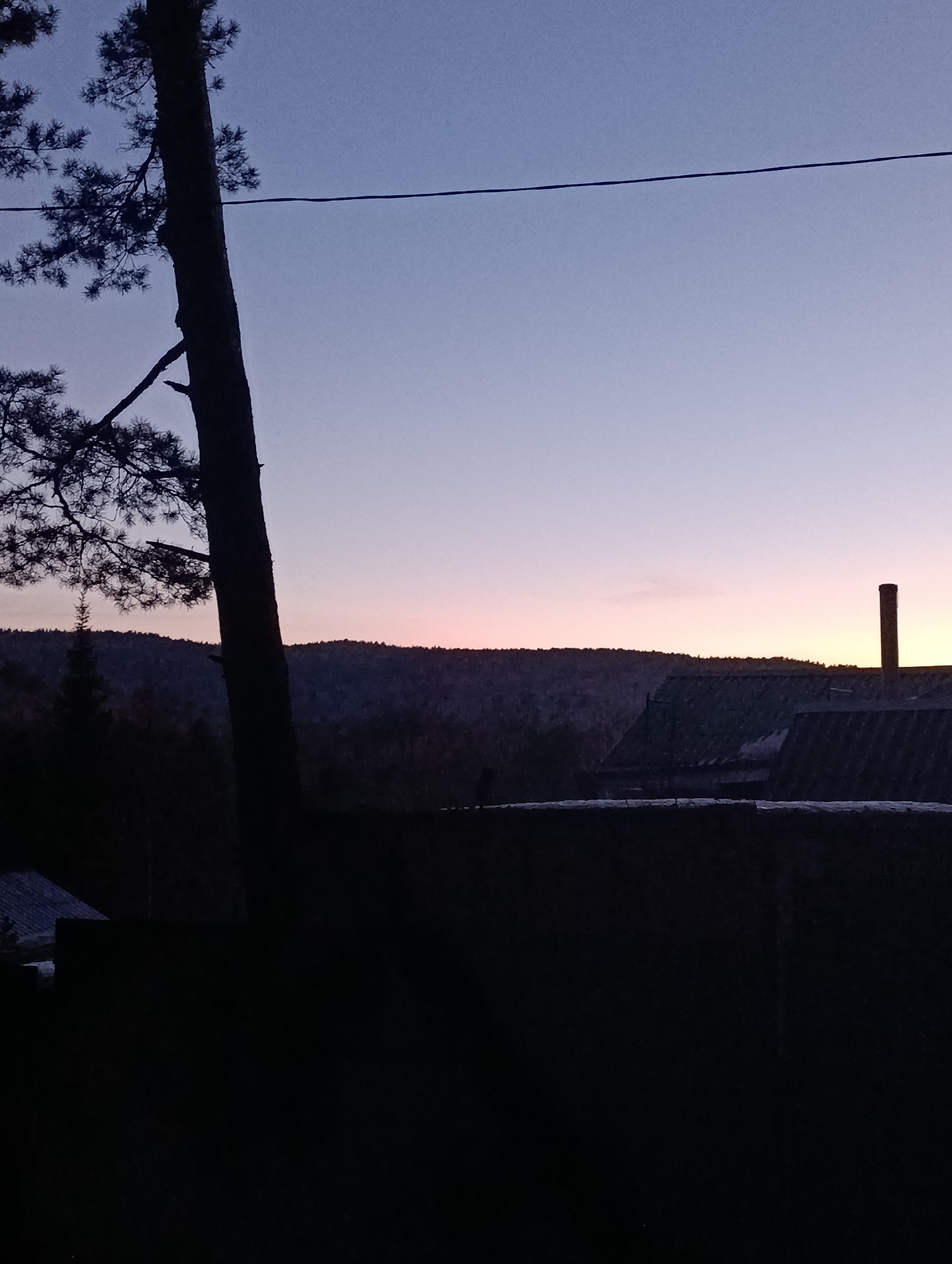 The image depicts a landscape at dusk in a rural area. In the foreground, there's a dark, silhouetted fence or barrier. To the left stands a tall, silhouetted tree with visible branches and foliage. Behind the fence, partially obscured, are silhouettes of buildings, including what appears to be a chimney. The background features a dark, forested hillside beneath a gradient sky transitioning from pale blue to faint orange near the horizon. A single power line stretches across the upper portion of the frame. It is difficult to confirm the location as Krasnoyarsk, Russia without additional reference points.