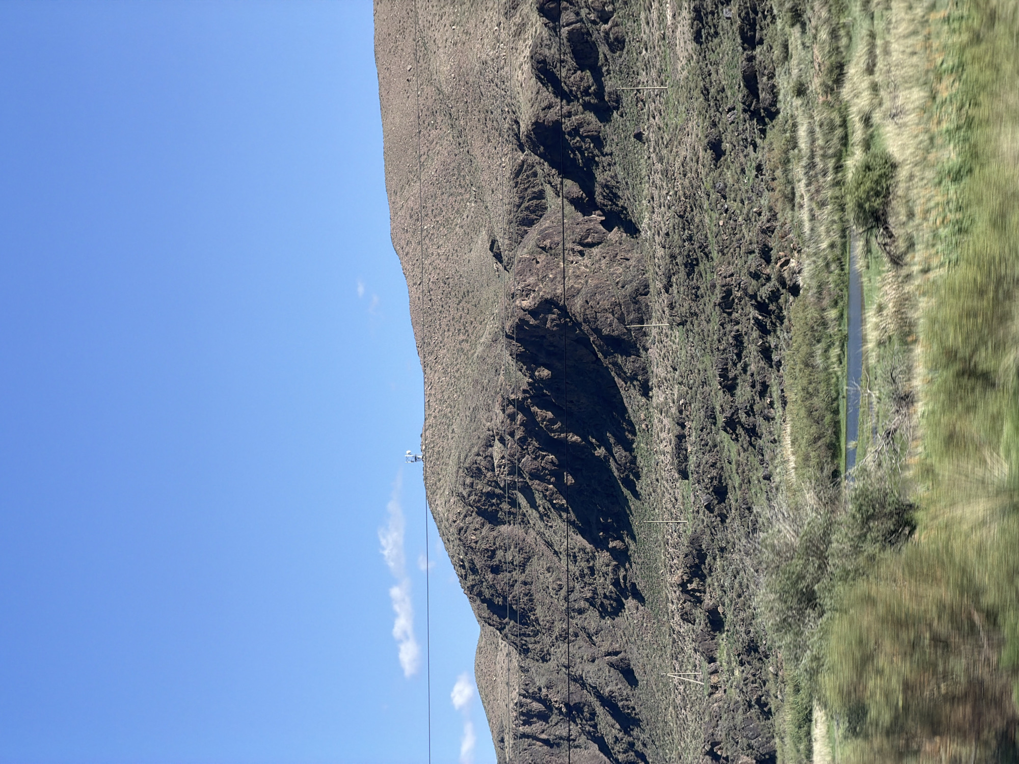 The image shows a vast, arid landscape under a clear blue sky. A rugged, rocky mountain dominates the middle ground, its slopes covered in sparse vegetation and dotted with dark, weathered rocks. Several utility poles and wires are visible traversing the mountainside, suggesting human infrastructure in this remote area. At the summit of the mountain, a small structure with antennas can be seen. In the foreground, a blurred stretch of green vegetation and a small body of water suggest a slightly more fertile area. This scene is likely located near Kok Moynok Eki, Kyrgyzstan, a region known for its mountainous terrain and natural beauty.
