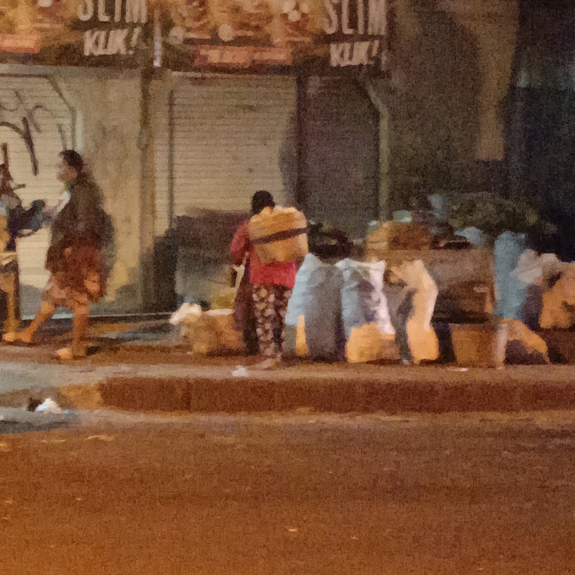 This image depicts a nighttime scene on a street in Salatiga, Indonesia, likely near a market or commercial area, as suggested by the blurred signage in the background that appears to advertise "SLIM KLIK". In the foreground, the street is visible with a sidewalk and a row of bundled goods, possibly merchandise or refuse, piled up.
Two individuals are the central focus. On the left, a person wearing a dark jacket and a patterned skirt is walking, carrying a bag over their shoulder. Their gait suggests they are moving away from the viewer. Further to the right, another person, seen from behind, is wearing a red top and patterned pants and has a large, light-brown bundle strapped to their back. This individual appears to be stationary or moving slowly. Between these two figures, several large, pale blue and white bundles are visible, some resting on the ground and others appearing to be partially covered.
The overall impression is that of a busy, perhaps informal, commercial or logistical operation taking place after dark, with individuals managing goods in what appears to be an urban setting. The lighting is dim, with some artificial light illuminating the scene from an unseen source, creating a gritty, realistic atmosphere. The pavement is wet, reflecting some of the light, and the background exhibits the texture of buildings or stalls.