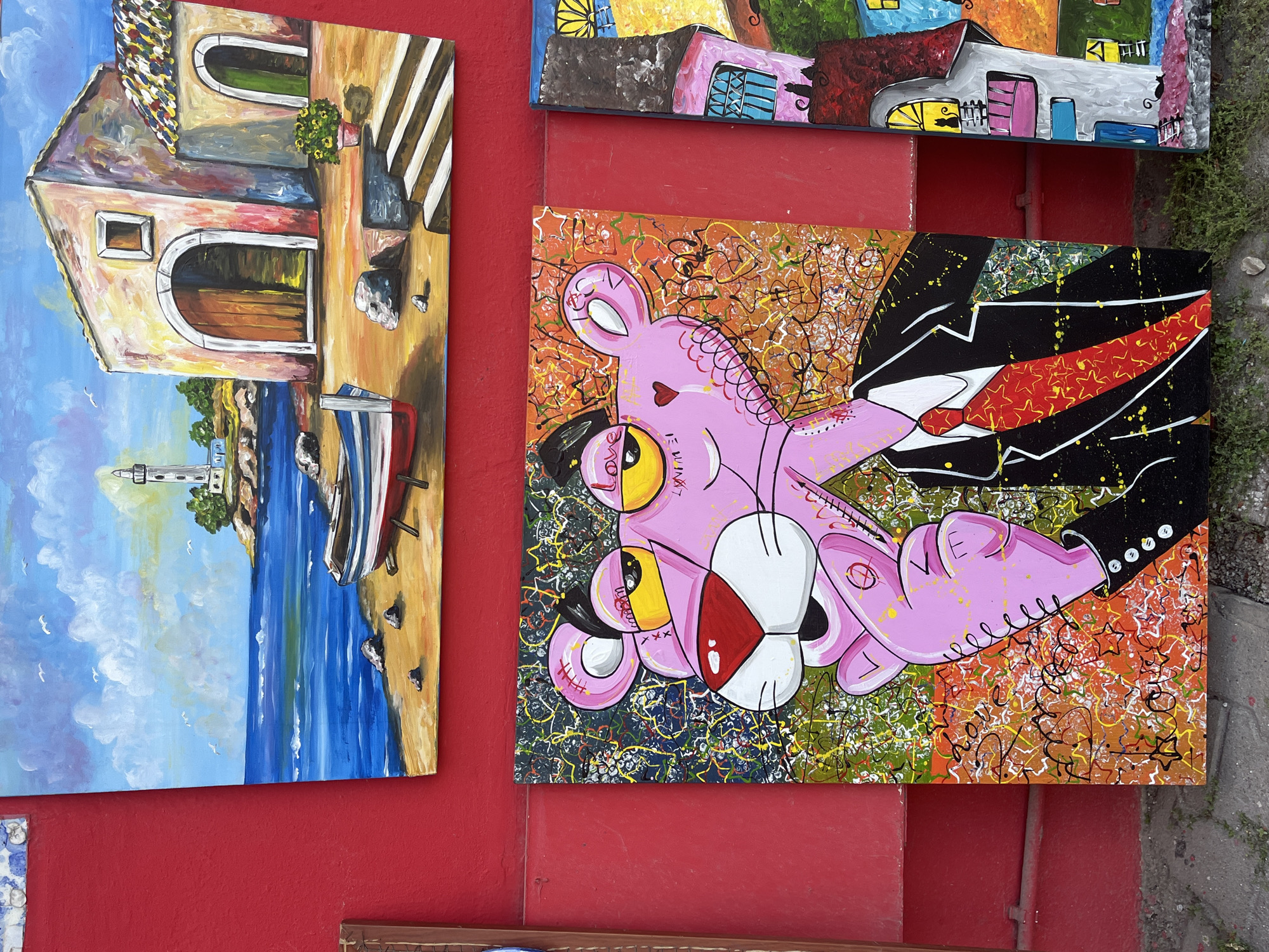 The image shows a display of paintings on a red wall in what appears to be an outdoor setting.
On the left, the painting depicts a coastal scene with a building, a lighthouse, a boat, and the sea.
Next to it is a painting featuring a stylized Pink Panther character wearing a suit. The background is a mix of colors and abstract patterns.
Other paintings are partially visible above and below these. The overall context is likely a street art display or an outdoor art market.
The location being Alanya, Turkey, is based on the general feel of the architectural and artistic style often found in tourist locations. The red painted wall resembles architecture that is common in Mediterranean tourist locations, including Turkey.