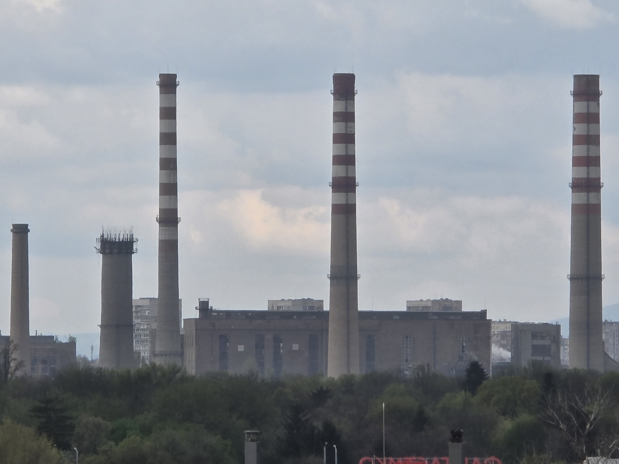 The image shows a panorama of a power plant in Sofia, Bulgaria. Several tall industrial chimneys dominate the scene. Some chimneys have a red and white striped pattern, while others are plain. The chimneys are located behind a line of trees. Behind the power plant and trees, there are buildings. The sky is cloudy and overcast.