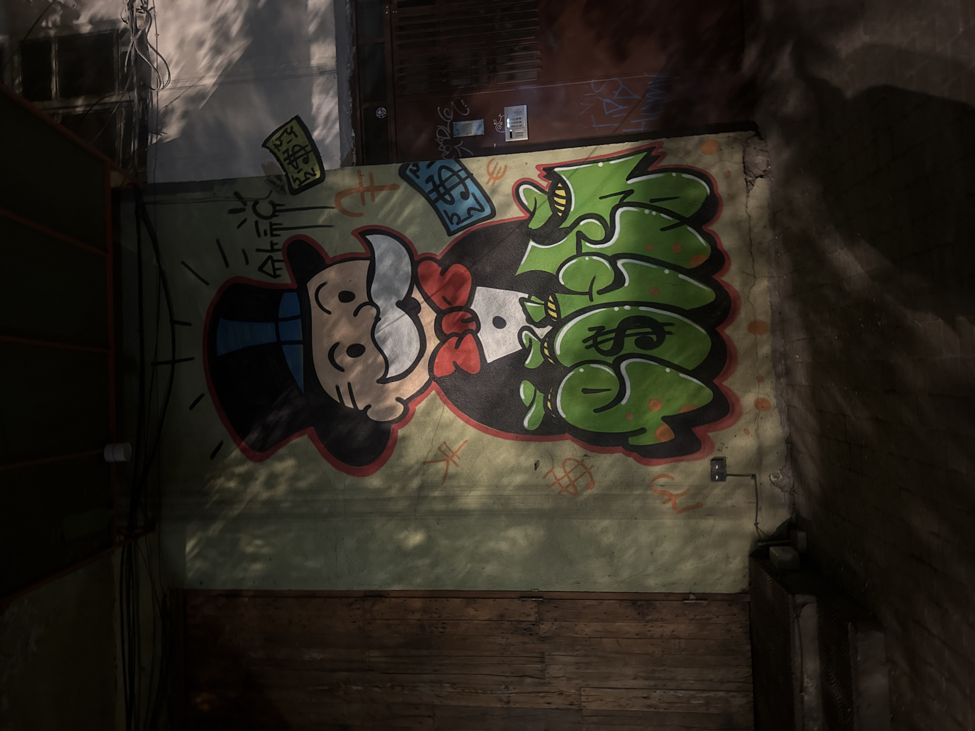 A horizontal medium shot taken at night shows a mural painted on a wall in Yerevan, Armenia. The mural depicts a stylized, cartoonish figure resembling the Monopoly man, rendered in a graffiti style. The figure has a large, exaggerated mustache and is wearing a top hat and a red bow tie. He is holding a stack of money, and his body is partially obscured by piles of green money bags, each marked with a dollar sign. The background of the mural is a light yellow color.
Above and around the figure, there are depictions of money symbols, including dollar signs and stacks of cash. The mural appears to be painted on a rough, textured wall that is slightly damaged in places.
The lighting in the photo is uneven, with strong shadows cast across the wall. Above the mural, some wires and other urban detritus can be seen. Below the mural, there is a wooden surface that appears to be part of a fence or platform. To the far right and left of the mural, there is a dark shadowy area.