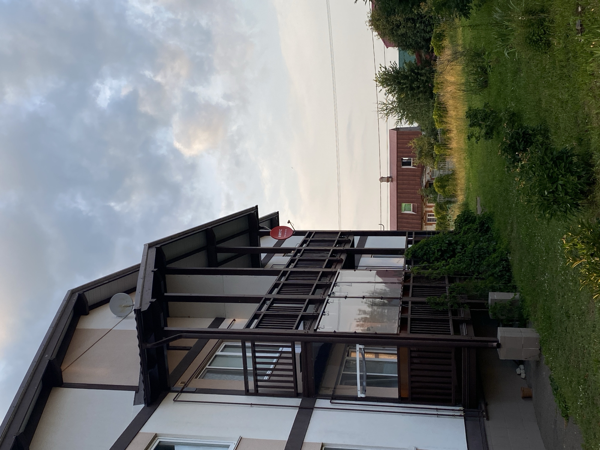 This is a medium-shot, exterior, eye-level, vertical photograph taken in Kuyuki, Russia, showing the side of a two-story building with a balcony and an overgrown yard. The building, painted in off-white and brown, has a prominent dark brown balcony on the second floor. The balcony has a wooden railing and is covered by a dark brown roof supported by wooden posts. Below the balcony, clear plastic sheeting is visible, and climbing vines are growing up the side of the building and the balcony supports. A satellite dish is mounted on the side of the building above the balcony, and a red circular sign with white text, possibly a logo, hangs from the balcony roof.
In the background, across a grassy yard with various plants and bushes, are other residential buildings. One building in the middle ground has a distinctive reddish-brown metal roof and appears to be a single-story structure with windows. Further in the background, more trees and houses are visible, suggesting a suburban or rural residential area. The sky above is partly cloudy with shades of gray and white, indicating overcast conditions or late afternoon light. The yard in the foreground is lush with green grass and interspersed with small bushes and flowering plants. The overall scene depicts a residential property in a somewhat overgrown state, under a cloudy sky in Kuyuki, Russia.