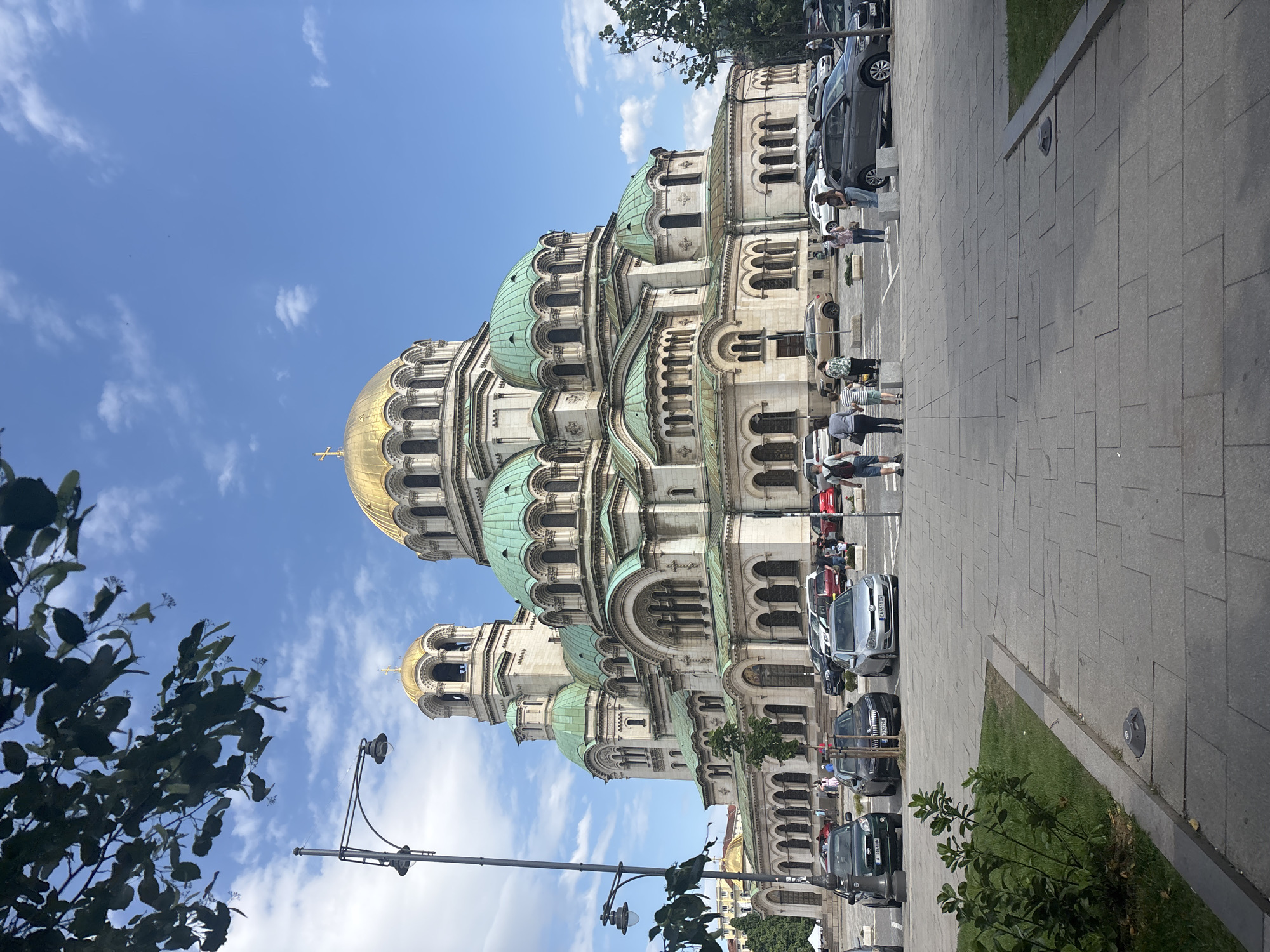 The image shows the Alexander Nevsky Cathedral in Sofia, Bulgaria. The cathedral has a golden dome and multiple green domes. The sky is blue with some clouds. Several people are walking on the sidewalk and some cars are parked along the street. Streetlights and trees are visible. The ground is paved with grey stone.