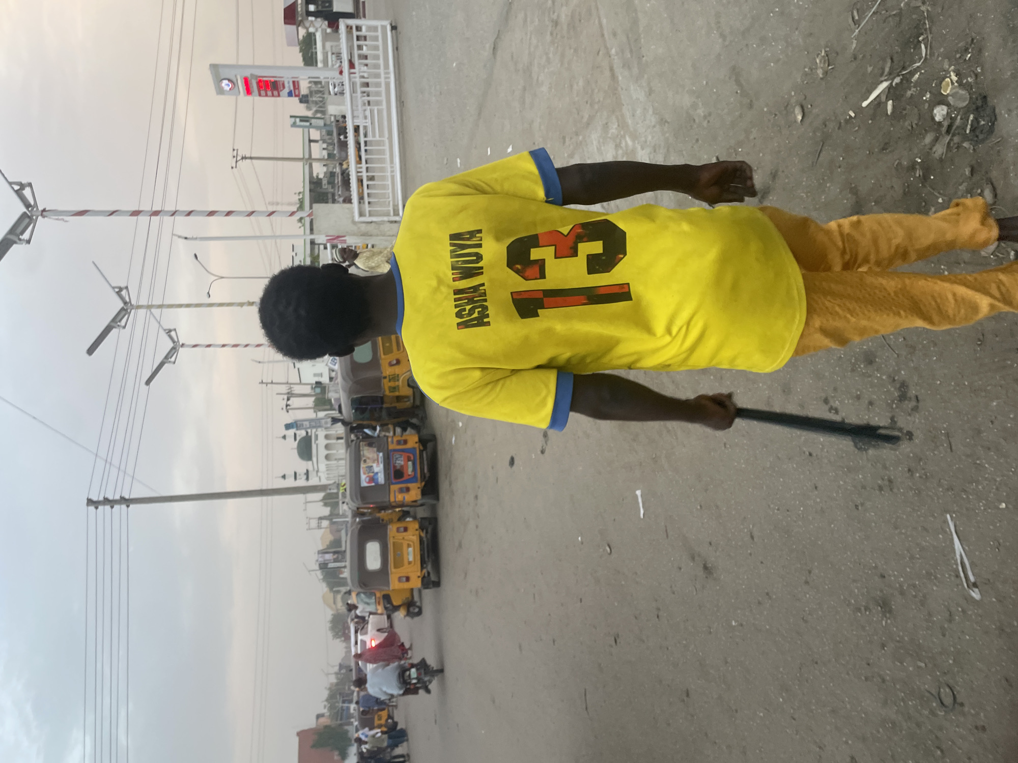 A medium shot shows a man wearing a yellow shirt with blue accents and the name "ASHA WUYA" and the number "13" on the back. He is walking on a paved road, holding a stick in his right hand. The road is cluttered with debris. In the background, there are several yellow auto rickshaws (tuk-tuks), some motorcycles, and other vehicles. A gas station is visible further in the distance. The image appears to be taken in a city in Unknown, Unknown.