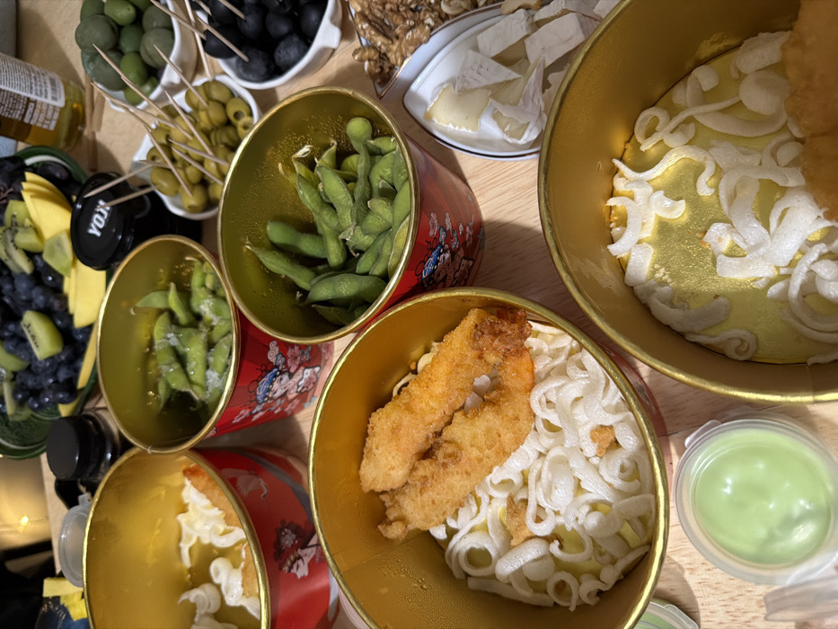 The image displays an indoor tabletop setting in Moscow, Russia, featuring a variety of food items arranged for consumption. The scene is illuminated by artificial light, including warm-toned fairy lights in the background.
The primary subjects are several cylindrical, red-and-gold containers, decorated with stylized Chinese opera figures. One container holds white, wavy noodles topped with two deep-fried, breaded shrimp. Two other identical containers are filled with green edamame pods, some visibly speckled with salt. A fourth container, partially visible, also contains white, wavy noodles. A fifth container appears mostly empty with some remnants of noodles.
Accompanying these, a small, clear plastic cup with a lid contains a pale green dipping sauce. Multiple white ceramic dishes are present: one holding light green olives with toothpicks, another with mixed green and black olives with toothpicks, and a third with black olives. An elongated white dish contains peeled walnuts, and another holds several slices of a soft, light-colored cheese.
In the upper background, a dark green ceramic bowl is filled with dark grapes, sliced kiwi, and sliced mango. A small black jar labeled "YOX" in white capital letters is visible near the fruit bowl. Partially obscured bottles, one clear glass containing a light liquid and another dark with a black cap, are also on the table. No people are visible, and no specific street or landmark in Moscow is discernible from the image.