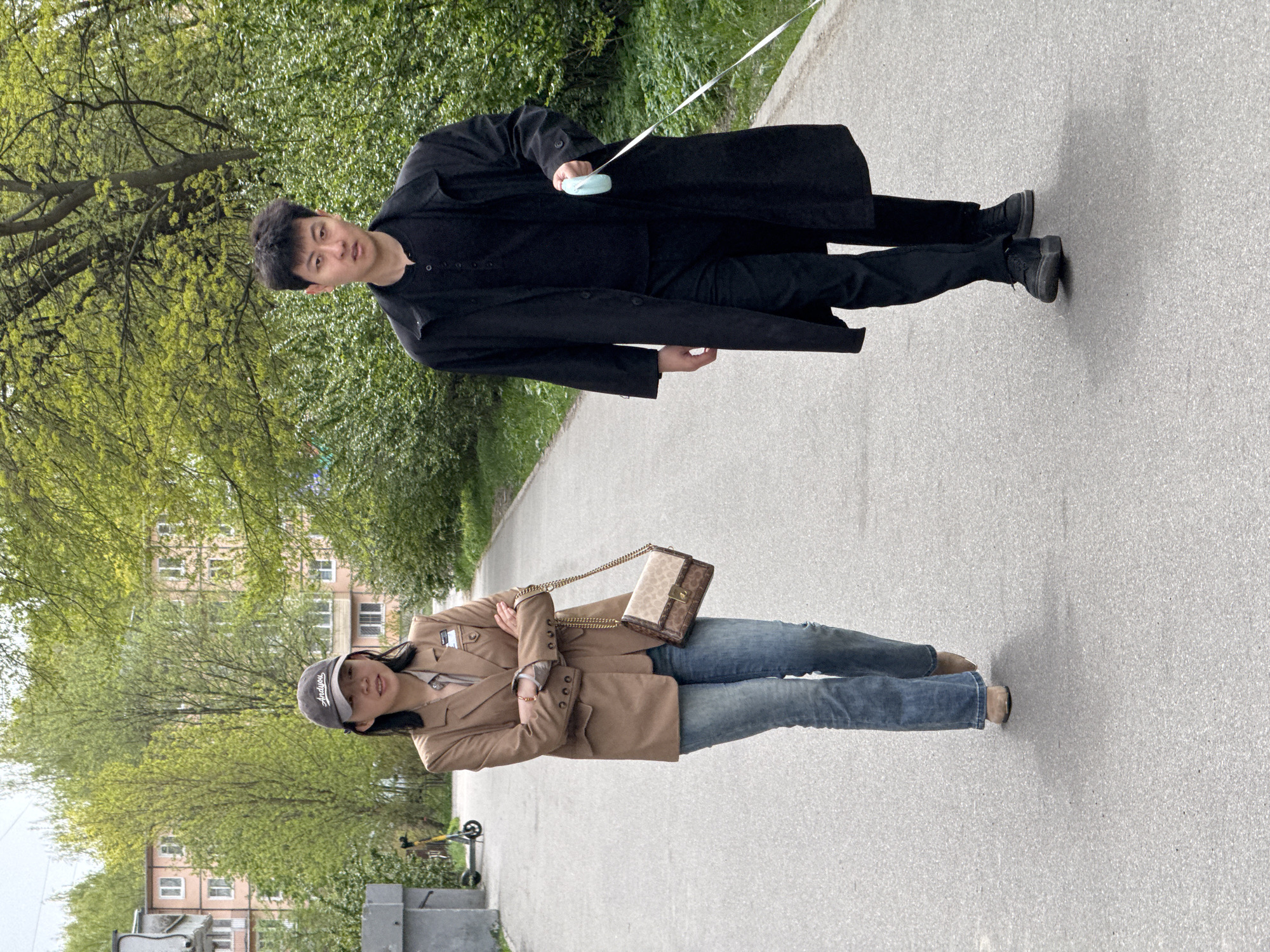 The image shows two people on a paved pathway in an urban environment. A person wearing a black coat, shirt, and pants is holding a white leash with a blue object at the end. They are walking toward the top of the frame. Below them, a second person is wearing a brown blazer, blue jeans, and a gray baseball cap with a brown purse hanging from their shoulder. They have their arms crossed. Behind them, trees, buildings, and a parked scooter are visible. Based on the architectural style and available information, the location is possibly in Saint Petersburg, Russia.