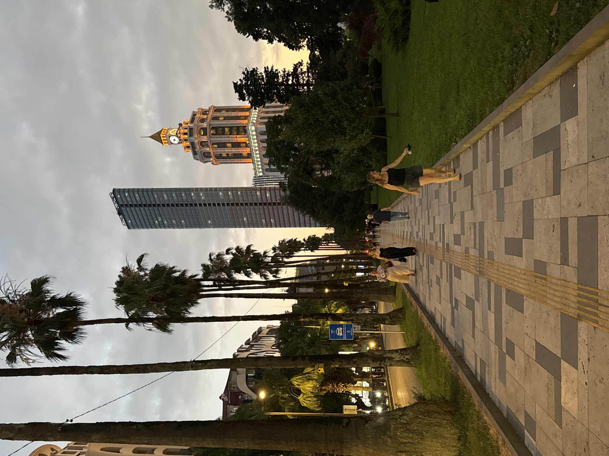 In Batumi, Georgia, on a wide, paved pedestrian walkway lined with palm trees, several people stroll as dusk settles. The walkway features a pattern of grey and beige square tiles, with a tactile yellow warning surface for the visually impaired running alongside it. A street with cars and streetlights is visible to the left, bordered by buildings and more palm trees. In the background, two tall modern buildings dominate the skyline: a slender, dark glass skyscraper and a distinctive ornate tower with illuminated windows and a clock face, suggesting a public square or boulevard. The sky is overcast with hints of sunset colors near the horizon. A woman in a black tank top and grey shorts walks towards the camera, holding a blue object. Another woman in a cream-colored dress walks ahead of her, followed by a man in a blue shirt and jeans.