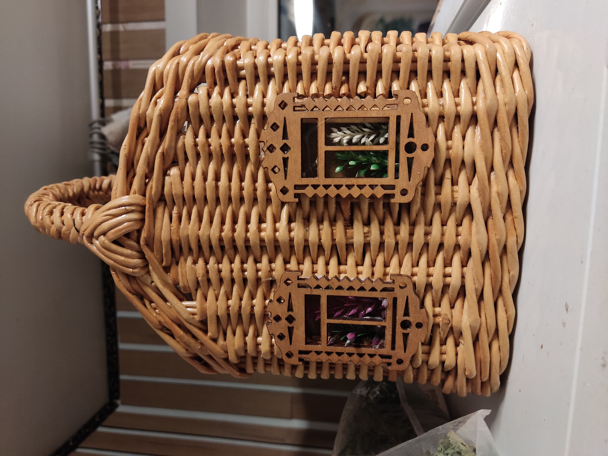 A close-up, eye-level, medium shot captures a woven wicker basket shaped like a small house, placed on a white shelf. The basket is light brown and features a thick, braided handle at the top. Two decorative wooden window frames, intricately carved with geometric patterns, are attached to the front of the basket. Through these frames, colorful artificial plants are visible. The basket is positioned in the foreground, with a blurred background that includes vertical striped wallpaper and other objects on shelves. The lighting is soft, highlighting the texture of the wicker. The specific location in the city of Noginsk, Russia cannot be determined from the image alone.