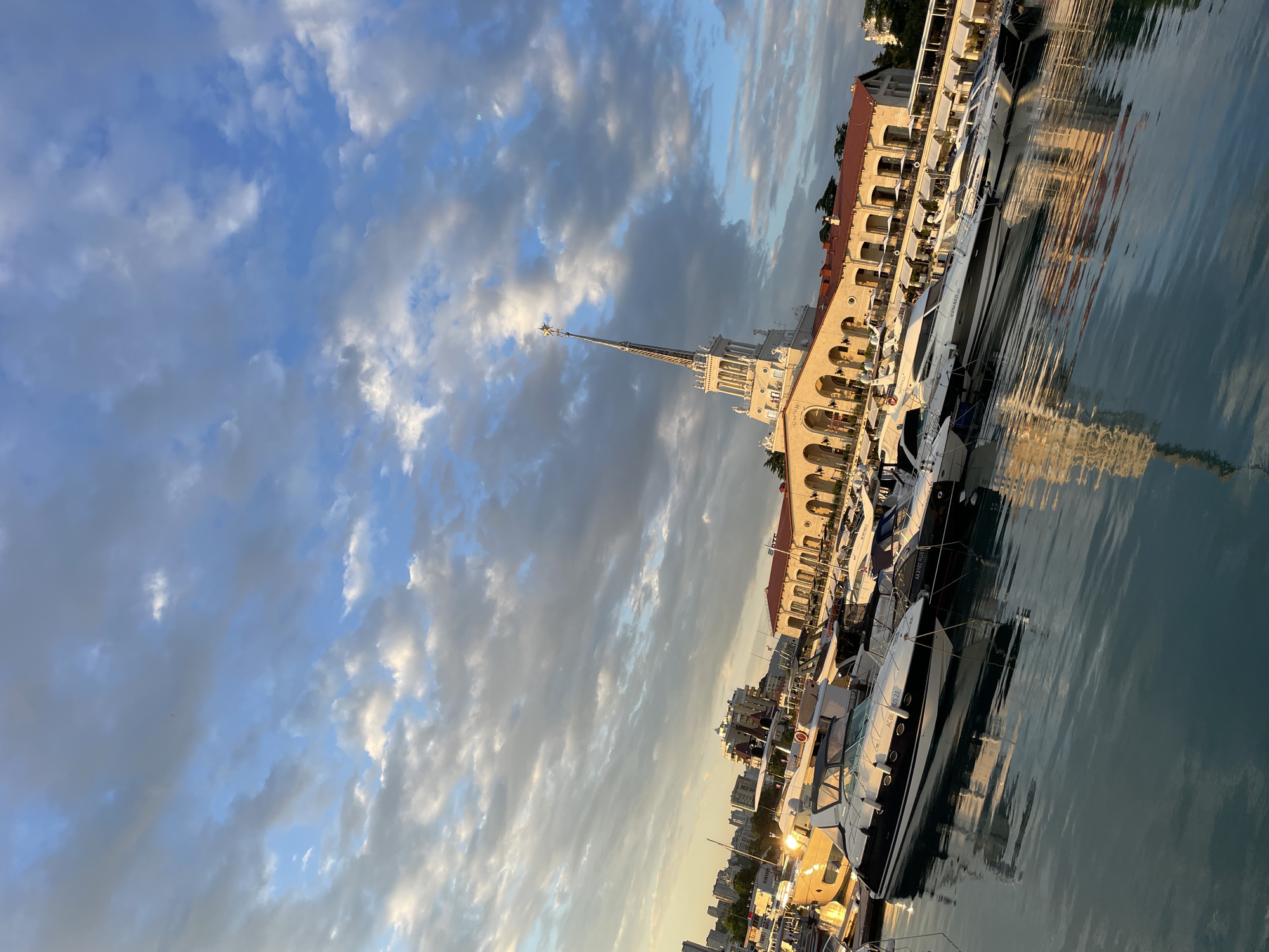 This image captures a scenic view of a harbor in Sochi, Russia, likely during the late afternoon or early evening. The sky is a dramatic canvas of blues and grays, with wispy white clouds catching the warm, golden light of the setting sun. The water is calm, reflecting the sky and the illuminated buildings and yachts docked along the quay.
The main architectural subject is a grand, cream-colored building with a distinctive tall spire topped with a star. This building features arched windows and balconies, suggesting a historical or classical style. Adjacent to this, several modern yachts of varying sizes are moored. Some have sleek, dark hulls, while others are lighter in color. Their reflections are clearly visible in the still water, creating an almost mirror-like effect.
In the background, more urban structures can be seen, indicating a developed coastal city. The overall atmosphere is one of tranquility and elegance, with the soft, warm light casting a serene mood over the scene. The image is oriented vertically, with the sky occupying the upper left portion and the harbor, buildings, and boats filling the rest of the frame. There is no visible activity of people in the frame, adding to the peaceful ambiance. Some text is visible on the yachts, but it is too small to be legible.
