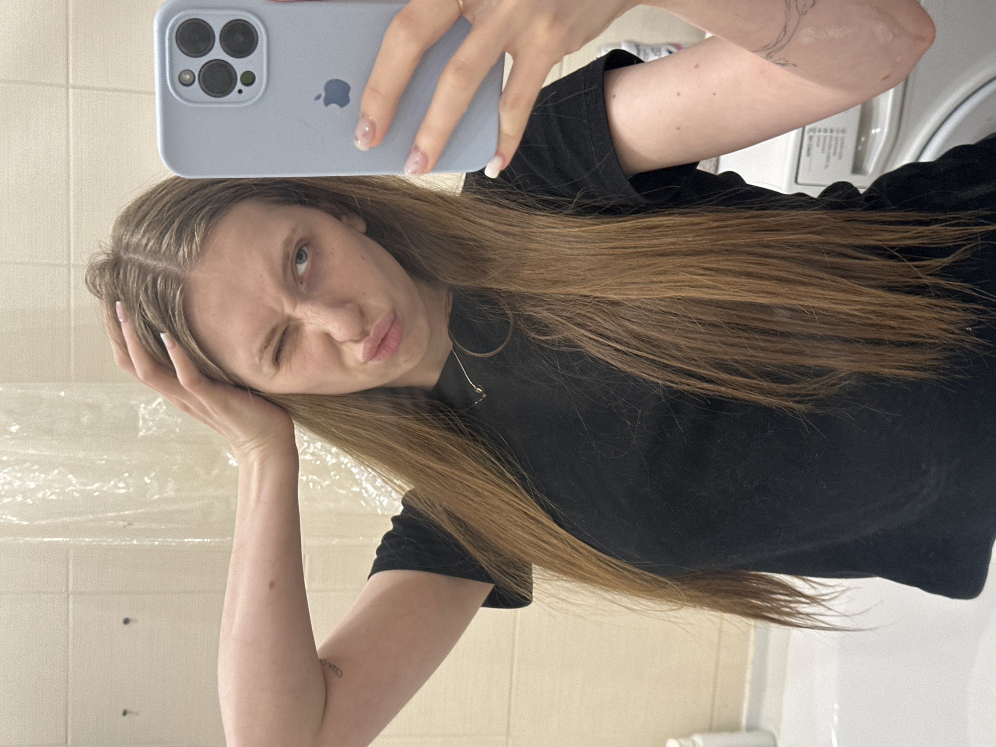 A young woman is taking a selfie in a bathroom. She holds a light blue iPhone with multiple camera lenses in her right hand, angled to capture her face and upper body. She has long, light brown hair and wears a black t-shirt. Her facial expression is playful, with one eye closed in a wink and her lips pursed. Her left hand is near her head, possibly adjusting her hair. The bathroom appears simple and functional with white tiled walls, a clear shower curtain, a washing machine, and standard bathroom fixtures. There are no discernible elements in the image to confirm the location as Chaikovsky, Russia.