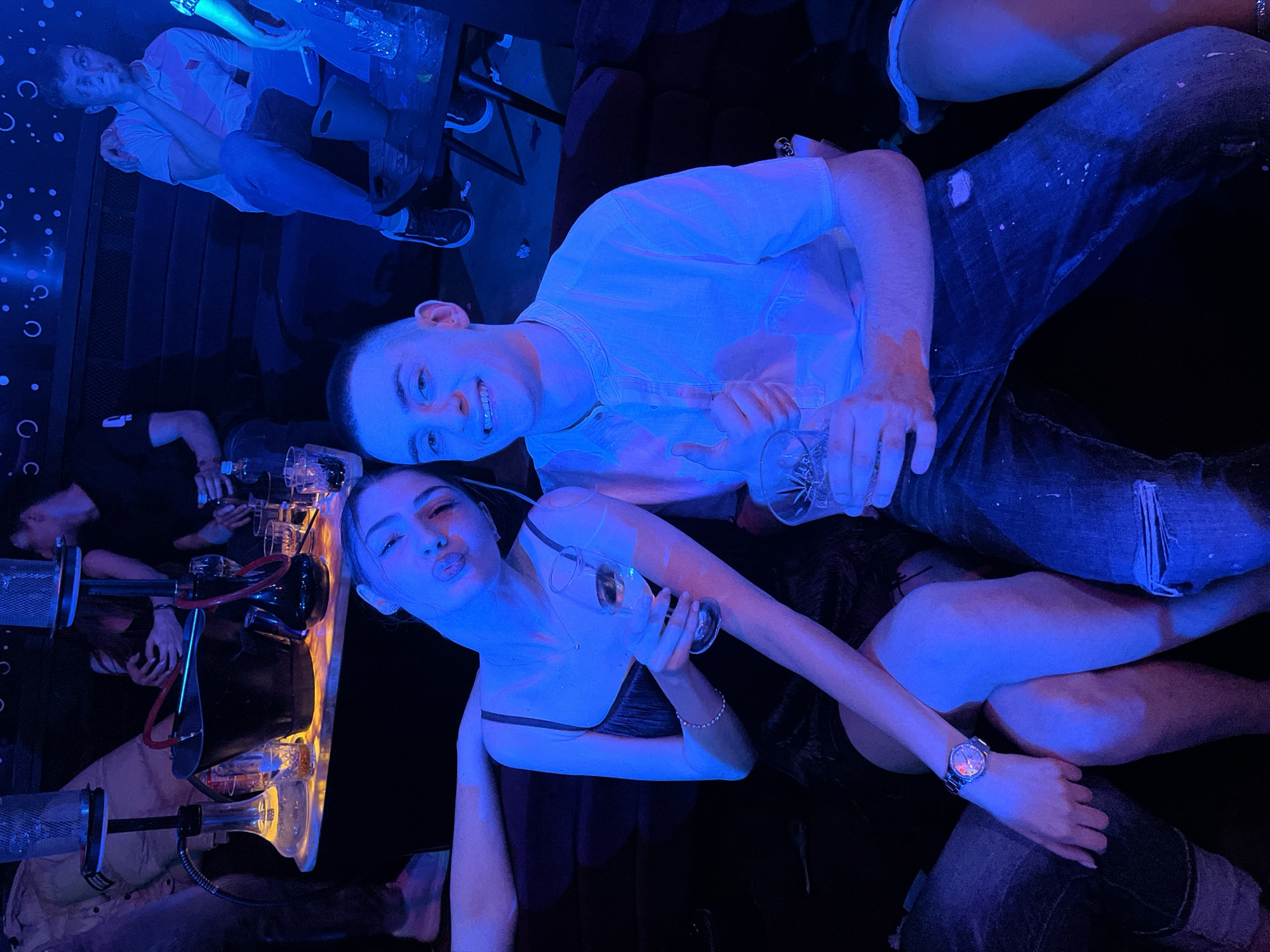 The scene depicts a group of people in what appears to be a nightclub or bar, possibly in Burgas, Bulgaria. The lighting is predominantly blue. Two individuals are in the foreground: a woman with long hair and a dark top is puckering her lips and holding a champagne glass. Beside her is a man wearing a light-colored collared shirt and ripped jeans, also holding a glass.
A table is positioned next to them, displaying what seems to be a hookah and various glasses. In the background, several other individuals are visible, some seated and others standing. One man is wearing a white shirt. Several figures are blurred.