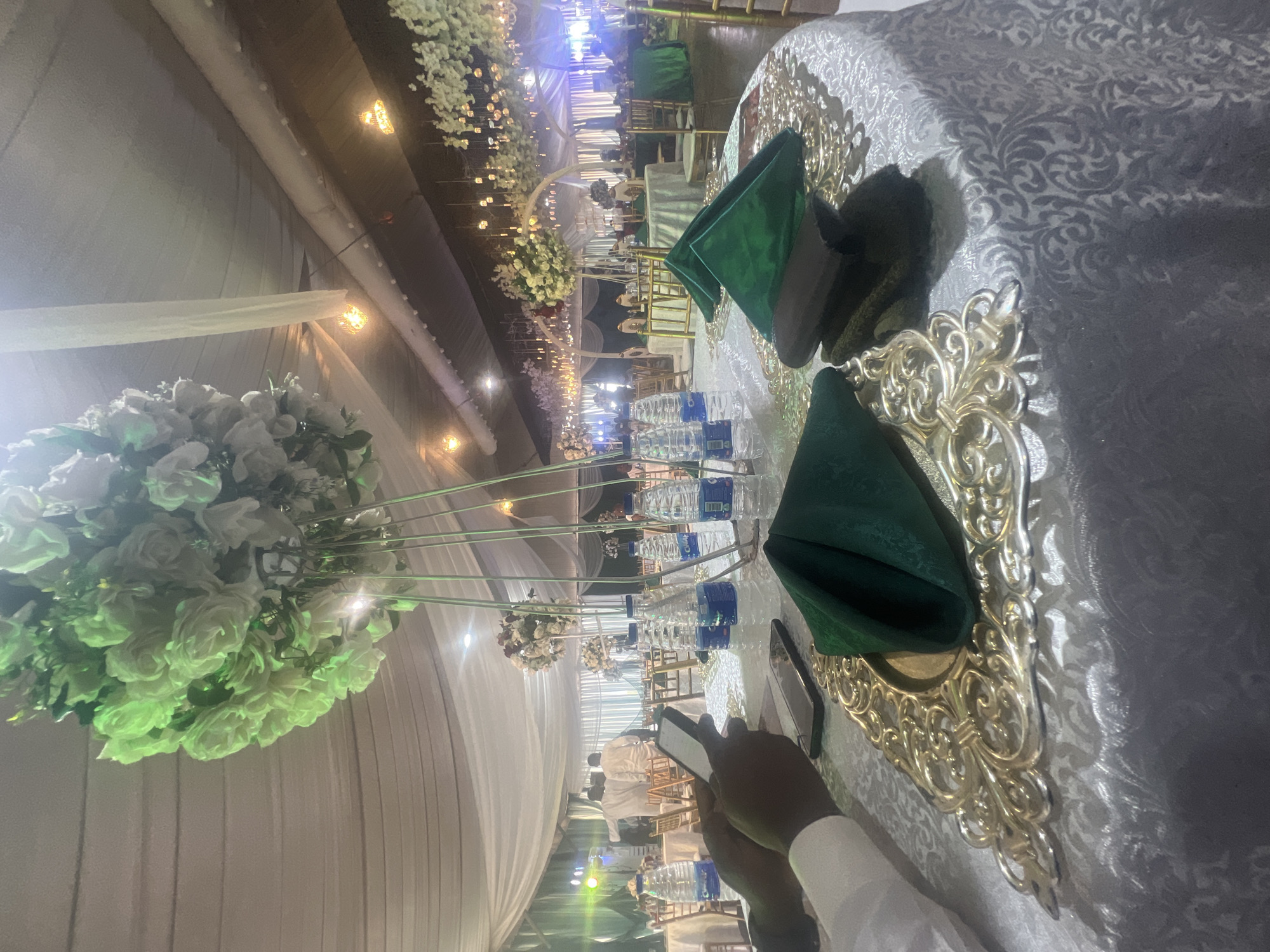 A long table at a wedding reception in Abuja, Nigeria. The table is covered with a patterned white tablecloth and decorated with gold chargers and green napkins. Water bottles are visible along the table. White floral arrangements are placed at intervals. Overhead, there is wedding decor with white flowers and string lights. A person is holding a smartphone, possibly taking a picture of the table. Other tables and guests are visible in the background.