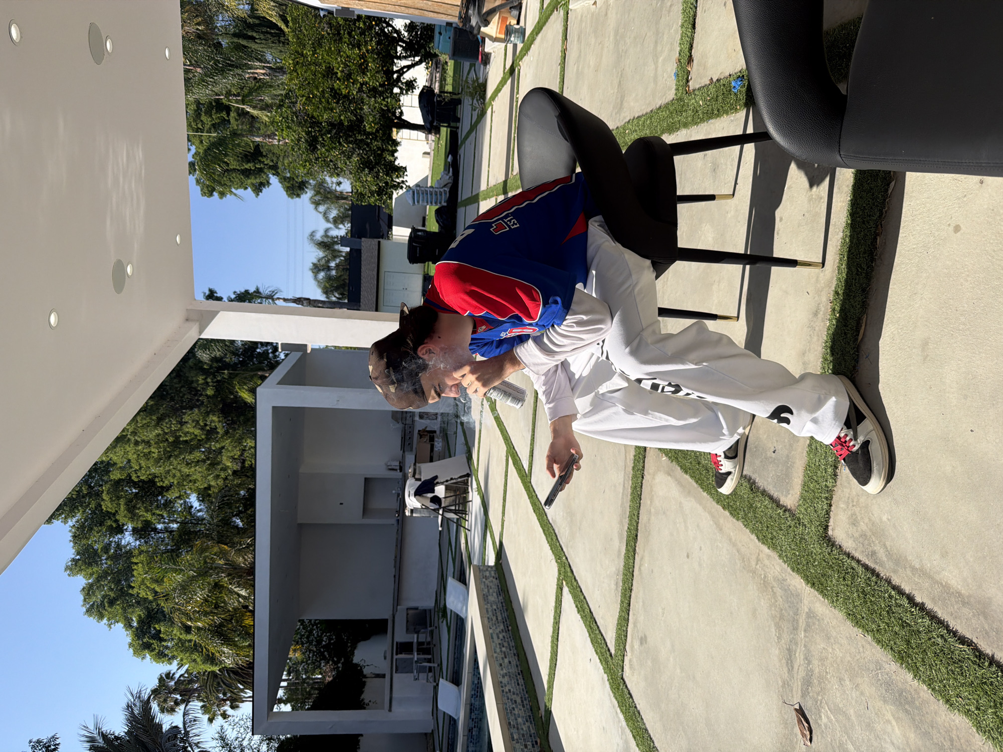 A person in a blue and red sports jersey, white pants, and sneakers is sitting on a black chair on a concrete patio in what appears to be a residential area in Los Angeles, United States of America. The person is holding a vaping device, and smoke is visible. The patio has artificial grass lines between the concrete slabs. In the background, there is a modern, minimalist-style house with trees and landscaping. Other patio furniture and outdoor structures are also visible. The time of day is likely midday, based on the position of the sun and the shadows cast.