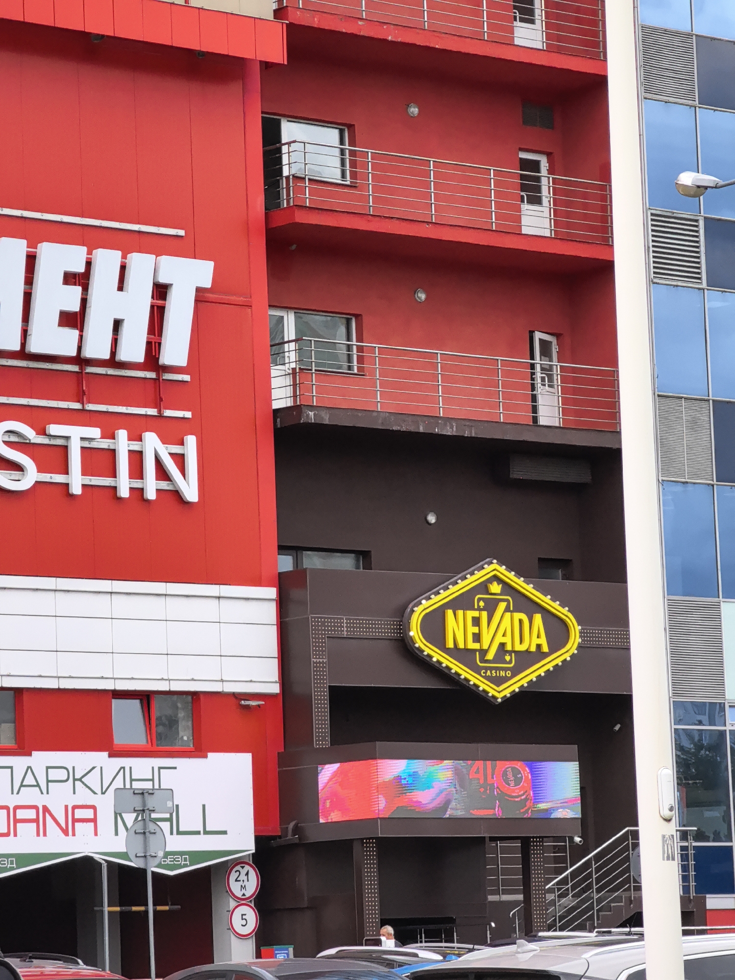 The image shows a close-up view of a multi-story building in an urban environment. The building features a prominent red facade with white lettering that appears to spell out "МЕНТ" and "STIN" on its upper levels. To the right, a modern building with blue glass windows and metallic louvers is visible.
At the lower level of the red building, a sign reads "ПАРКИНГ OANA MALL". Below this, there are entrances to a parking facility. Adjacent to the red building is a section with a dark brown facade, featuring a neon sign for "NEVADA CASINO". A large digital screen displaying vibrant abstract patterns and casino-related imagery is mounted below the casino sign.
The scene includes several vehicles parked in front of the building entrances, suggesting a public space like a shopping mall or entertainment complex. A traffic sign indicating a height restriction of 2.1 meters and a circular sign with the number 5 are also visible. The overall impression is of a commercial or entertainment district within a city.