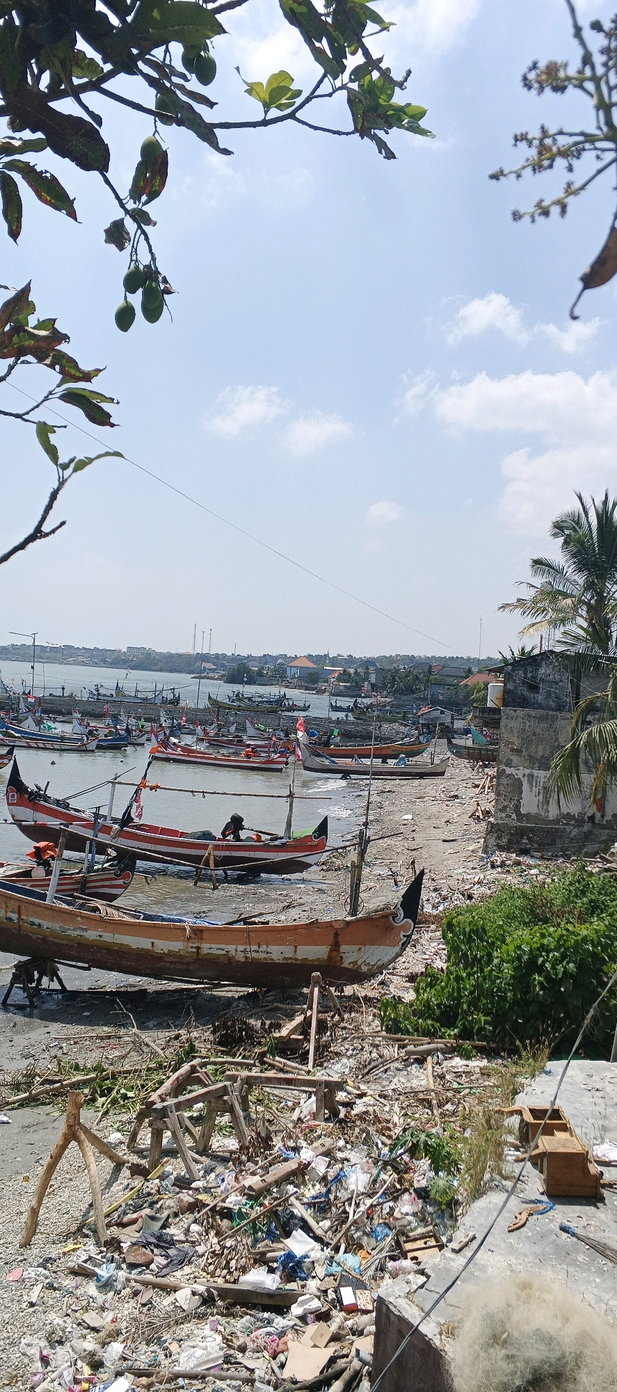The image depicts a coastal area with numerous traditional fishing boats docked on the shore and in the shallow water. The scene appears to be in Kandangsemangkon, Indonesia. In the foreground, there is a significant amount of debris and trash scattered across the beach, including plastic waste, wood, and vegetation. Several wooden structures, possibly for supporting boats, are also visible.
The boats themselves are varied in color and design, many featuring vibrant red and white stripes, and some with decorative elements. A few individuals are present, seemingly tending to the boats or engaged in activities related to fishing. The water is calm, and in the background, a town or village with houses and buildings can be seen along the coastline, with palm trees dotting the landscape. The sky is mostly clear with scattered white clouds. The overall impression is that of a working fishing harbor, albeit one affected by considerable litter.