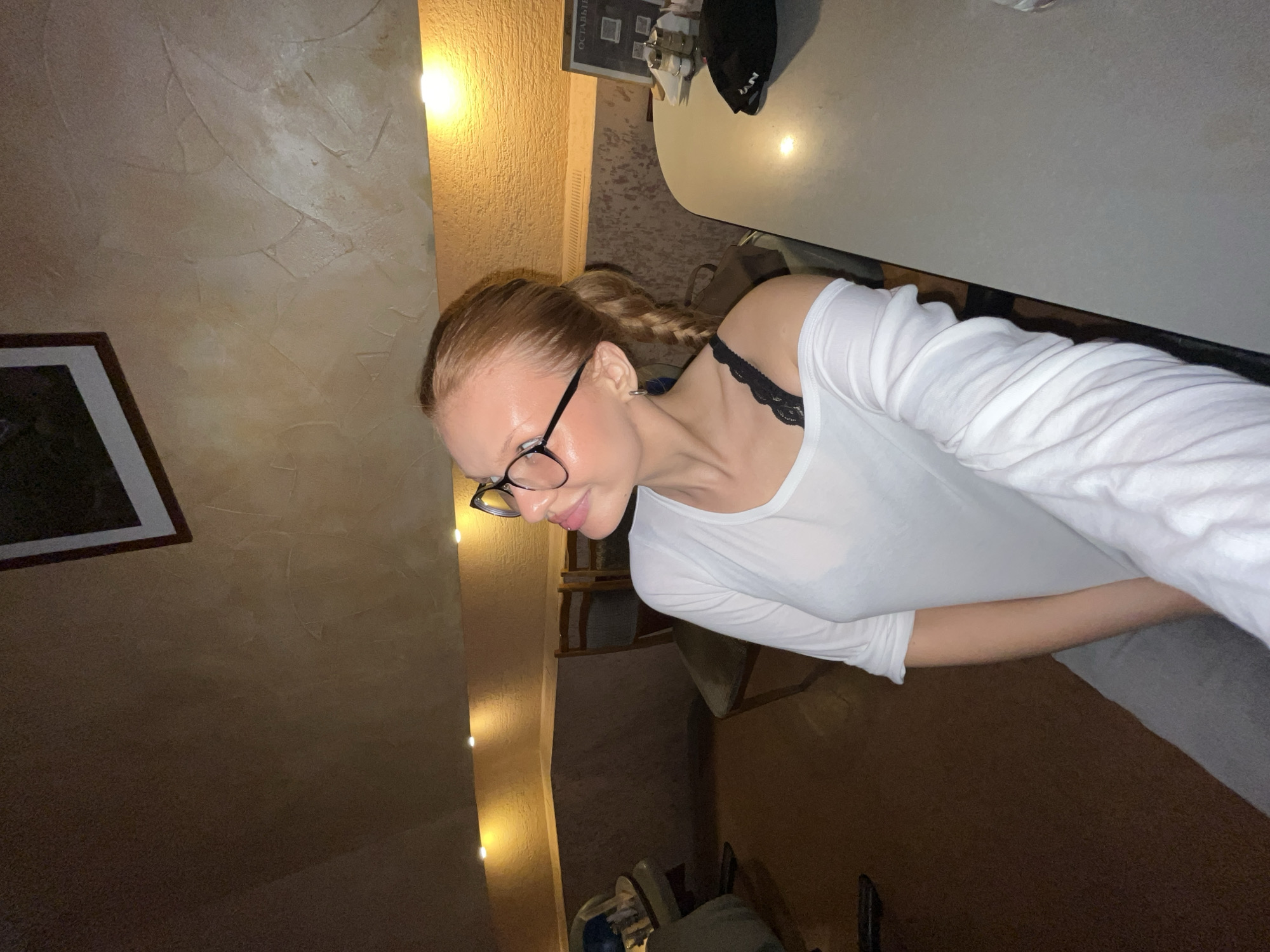 A woman with blonde hair styled in a braid and wearing glasses and a white shirt is pictured. She is seated at a table in what appears to be a restaurant with warm lighting and textured walls. In the background, there are chairs and a framed picture on the wall. The image is cropped to focus on the woman and her surroundings.