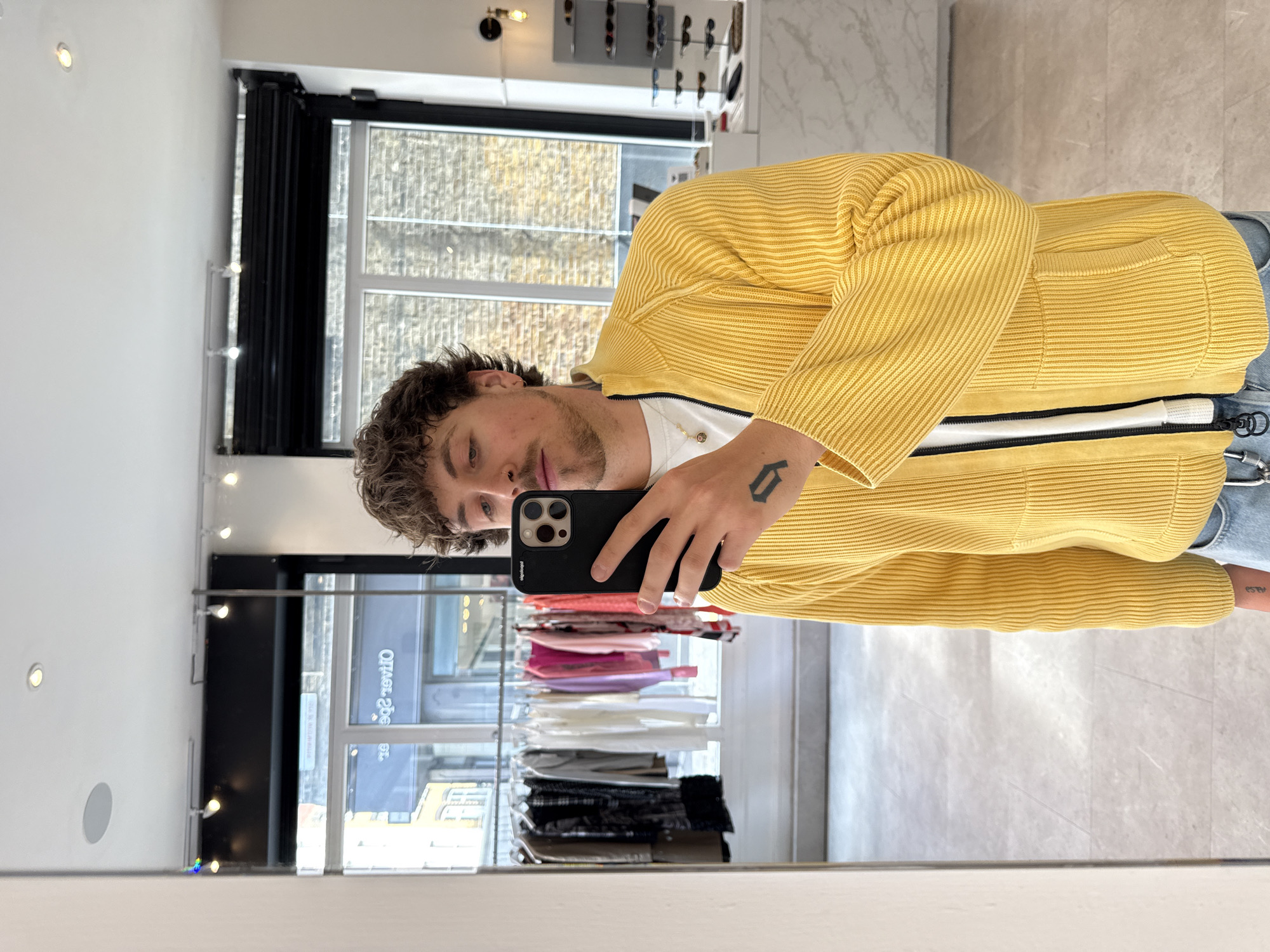 A man takes a mirror selfie in a clothing store in London, United Kingdom. He is wearing a yellow knit sweater. He is holding a black smartphone in his left hand to take the photo. A tattoo is visible on his hand. The reflection in the mirror shows clothing racks with various items. Outside the store, a building with a white brick facade is visible through the window.