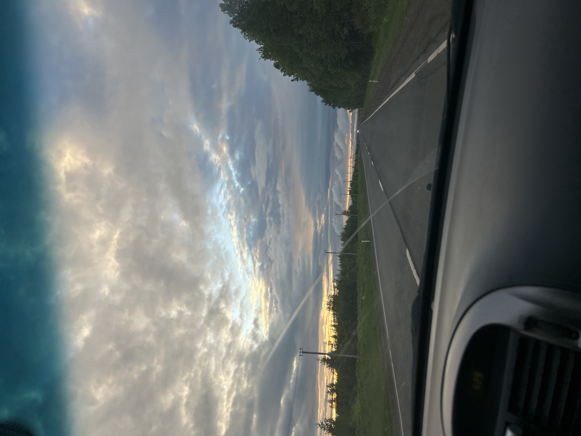 The image depicts a view from inside a car, looking out through the windshield. The car appears to be traveling on a road adjacent to a forested area. The sky is filled with clouds, some of which are illuminated by the setting sun. Telephone poles line the edge of the road. The location is Gorshki, Russia.