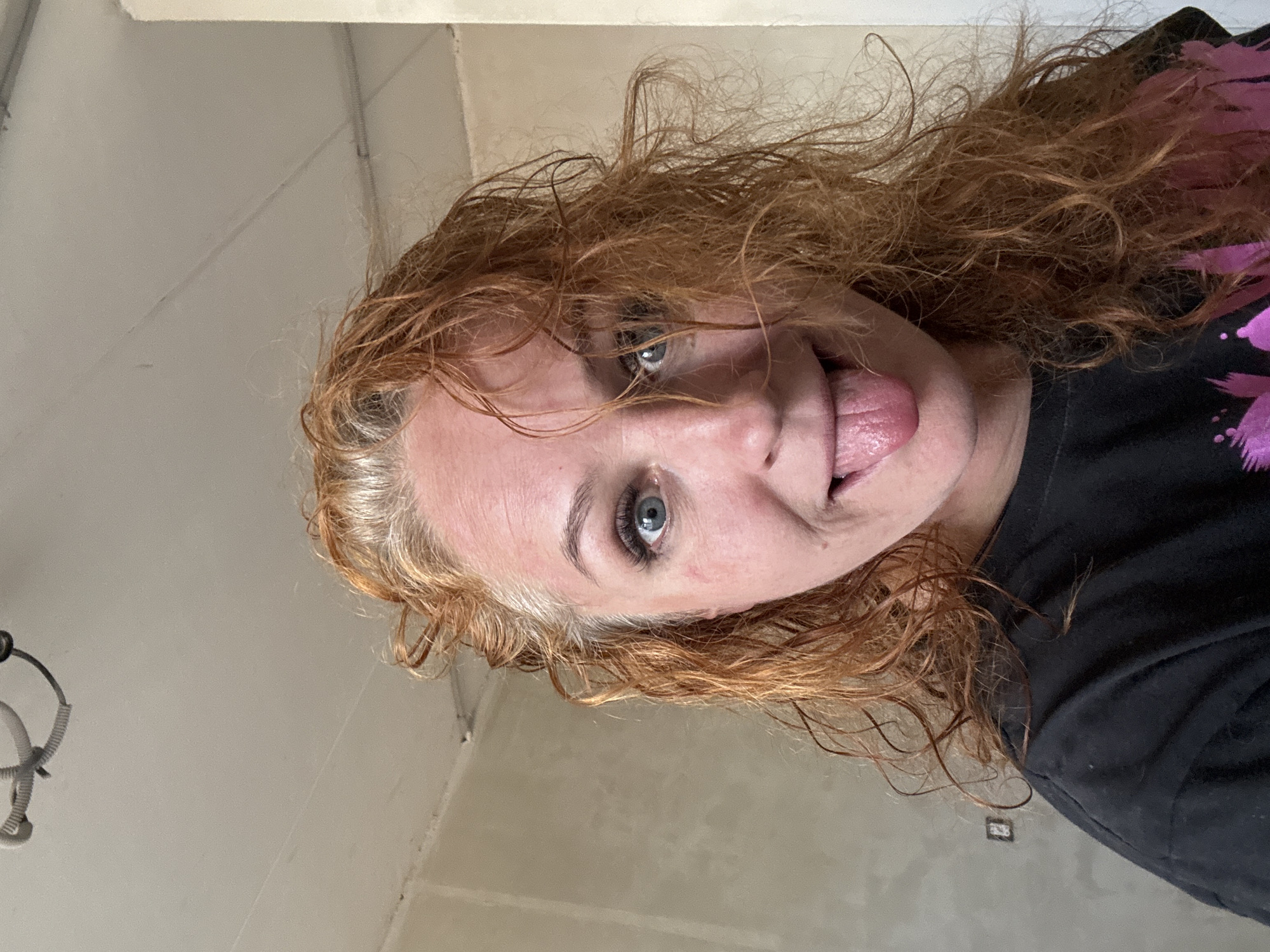 A selfie angled upward depicts a woman with light skin, blue eyes, and reddish-brown, curly hair sticking her tongue out. She is wearing a black t-shirt, partially visible in the lower right. The background consists of light-colored walls, including a textured wall and a white ceiling with exposed wiring. There is no evidence to suggest this image was taken in Chaikovsky, Russia.
