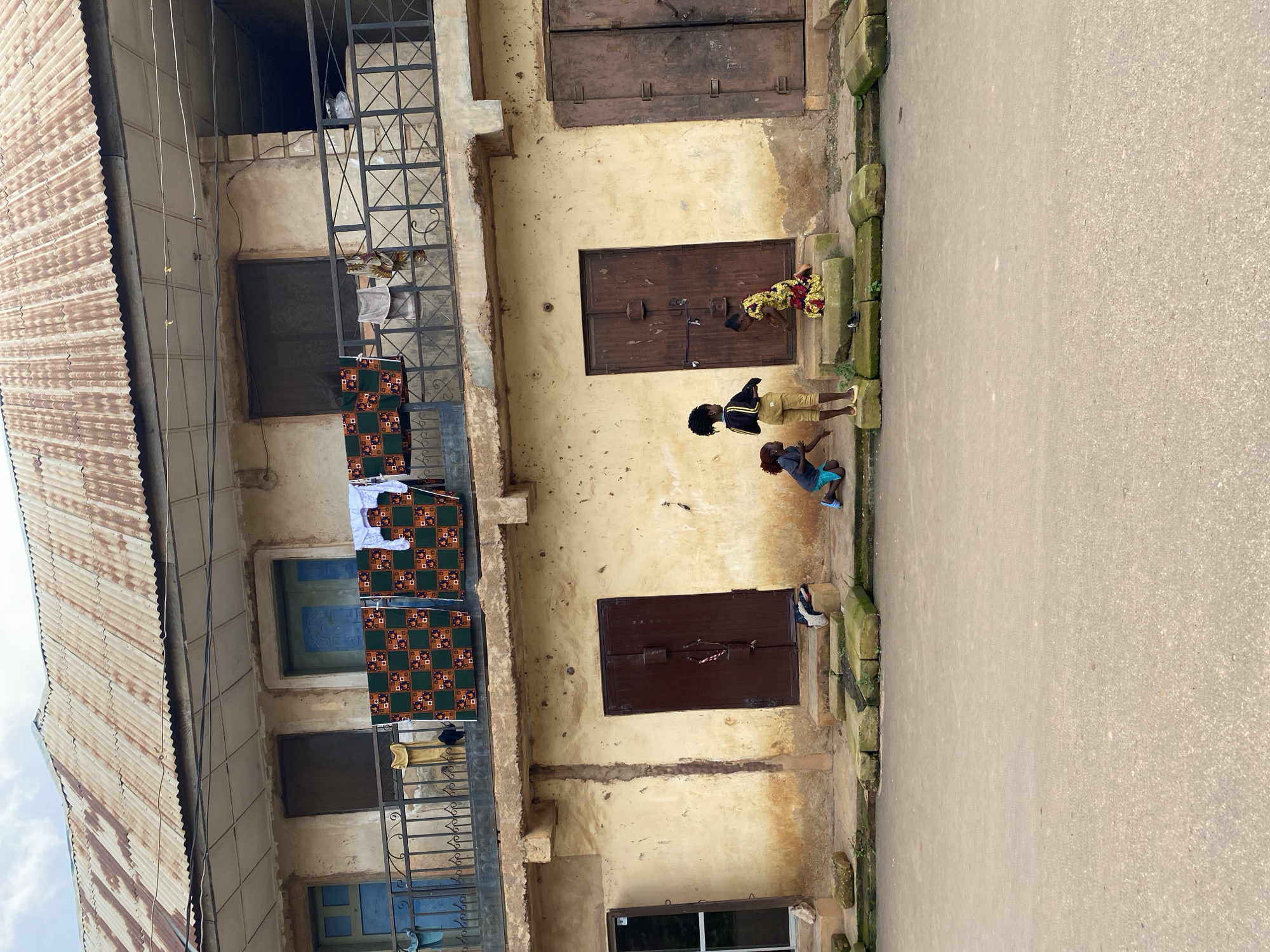 The image depicts a street scene in Gura Top, Nigeria, featuring a two-story building with a weathered, corrugated metal roof. The building's facade is constructed of roughly plastered, cream-colored walls, stained with age and possibly rain. On the upper level, a balcony with a black metal railing is visible, adorned with drying laundry, including colorful patterned cloths and white garments. Several windows and doors punctuate the building's facade, some painted in shades of blue.
At ground level, three children are interacting in front of the building. One child, wearing a yellow and black striped top and tan pants, stands near a dark brown double door. Another child, wearing a blue shirt and light blue shorts, is kneeling to the left of the standing child. A third child, dressed in a vibrant yellow and red patterned dress, is kneeling on the steps leading to the double doors. A dark brown double door with visible latches and a chain is centrally located. To the left of this door is a smaller, dark-framed window. To the right, another larger, weathered dark brown double door is present. The ground in front of the building consists of uneven, green-tinged stone blocks and a paved asphalt street. The overall scene suggests an outdoor, everyday setting within a residential area.
