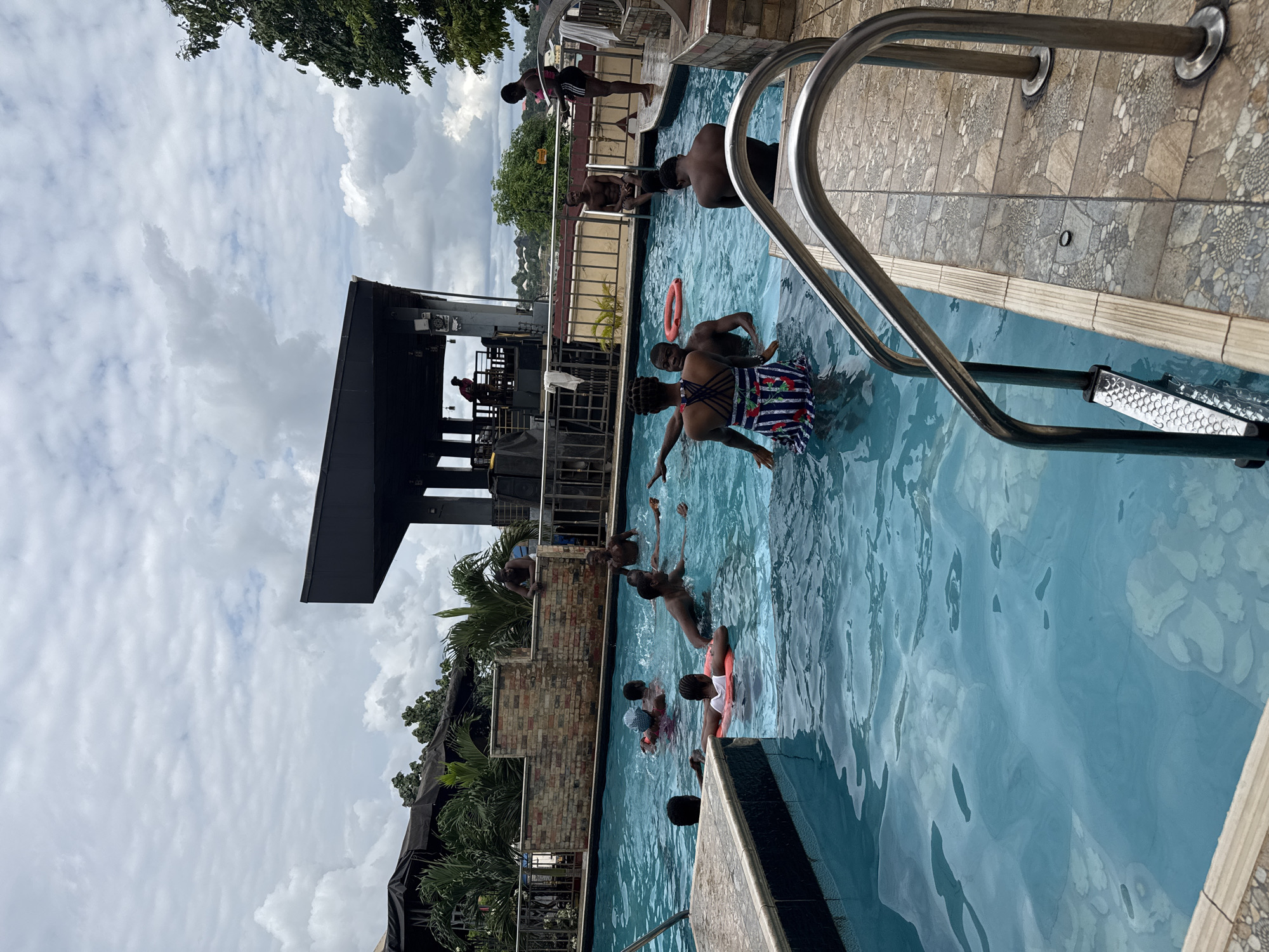 The image captures a lively scene at an outdoor swimming pool in Nkpor, Nigeria. Several people, predominantly young adults, are enjoying the water under a cloudy sky. The pool features a tiled edge and a prominent stainless steel ladder, suggesting a public or resort setting.
Several individuals are actively swimming or playing in the pool. In the foreground, a person wearing a patterned skirt is partially submerged, looking towards the camera. Another person in a red inner tube is visible further back. Other individuals can be seen in different parts of the pool, some interacting and others simply relaxing in the water. On the edge of the pool, a few people are standing or sitting, observing the activity. A structure that appears to be a bar or restaurant with a dark roof is visible in the background, with a few people near it. Palm trees and lush greenery surround the pool area, indicating a tropical or subtropical climate. The overall mood is one of recreation and enjoyment. The time of day appears to be afternoon, given the ambient light. There is no visible text in the image.