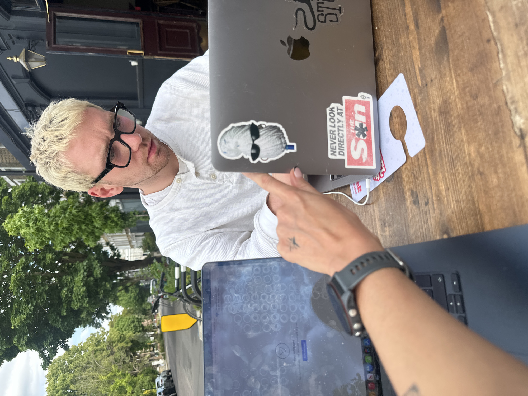 The image depicts a man with bleached hair and glasses, seated outdoors at a wooden table. He is wearing a white, long-sleeved, button-down shirt. The man is looking at a laptop computer that is being pointed at by a woman’s hand. The laptop has stickers on it, one of which reads "NEVER LOOK DIRECTLY AT THE Sun." Another laptop, possibly a tablet, is visible in the foreground, displaying messages. The woman's wrist with a watch and a star tattoo are also visible. Trees and what appears to be a street scene are visible in the background, indicating an outdoor setting, potentially a café or patio in London, United Kingdom. The building in the background appears to be a period building.