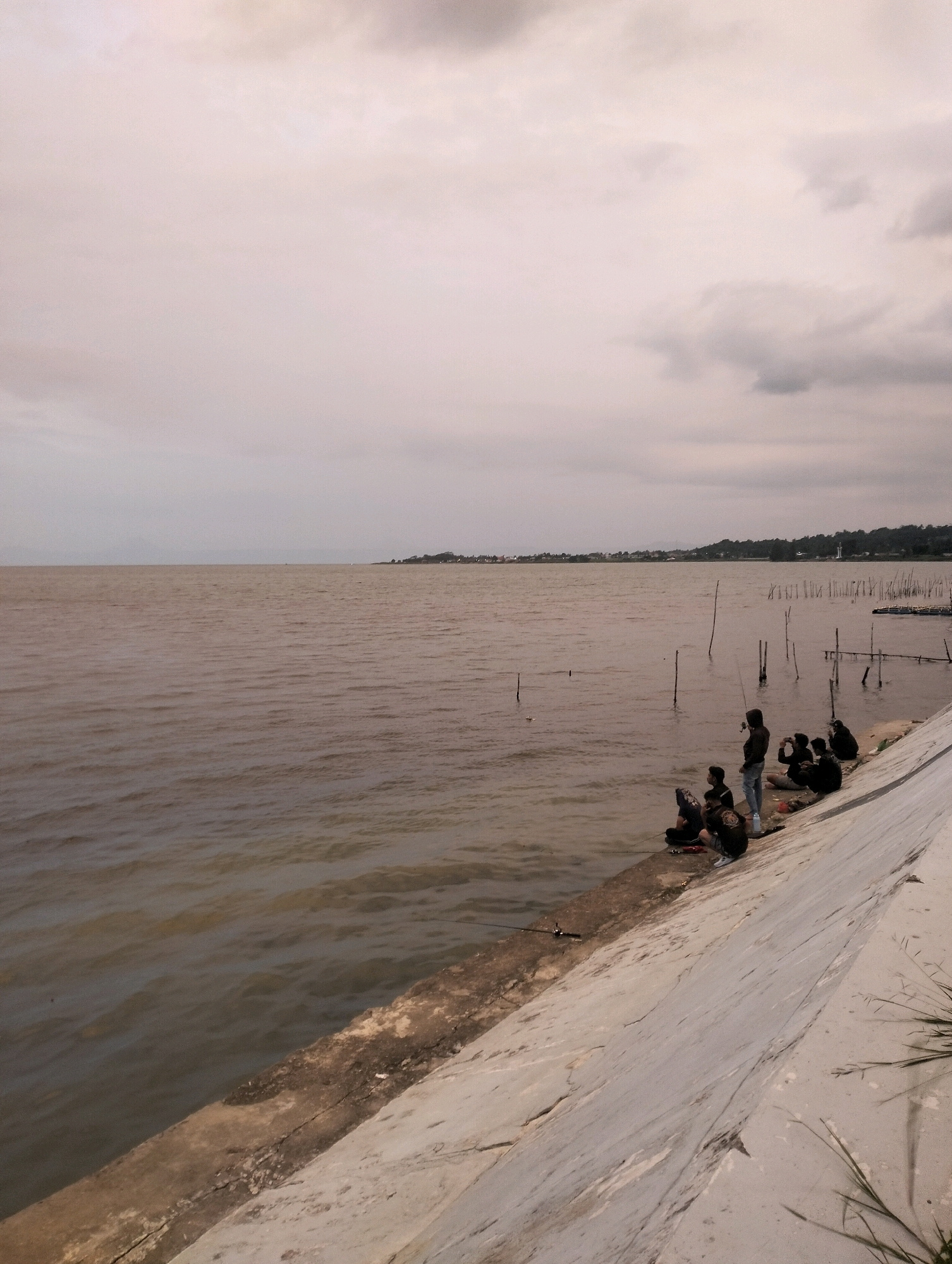 The image depicts a cloudy day by a large body of water, likely a lake or bay, with a village or town visible on the distant shore. In the foreground, a concrete embankment slopes down to the water's edge. A group of at least six individuals are seated or squatting along the embankment, engaged in fishing. They are mostly dressed in dark clothing. Some are holding fishing rods, while others appear to be waiting. The water is calm and has a slightly murky, brown-green hue. There are several wooden poles or stakes protruding from the water near the shore, possibly part of a fishing net or marker system. The sky is overcast with diffused light, suggesting a late afternoon or early morning scene. The environment appears to be natural, with some sparse vegetation visible on the embankment. Based on the context of Parsaoran I, Indonesia, this scene is likely a popular fishing spot.