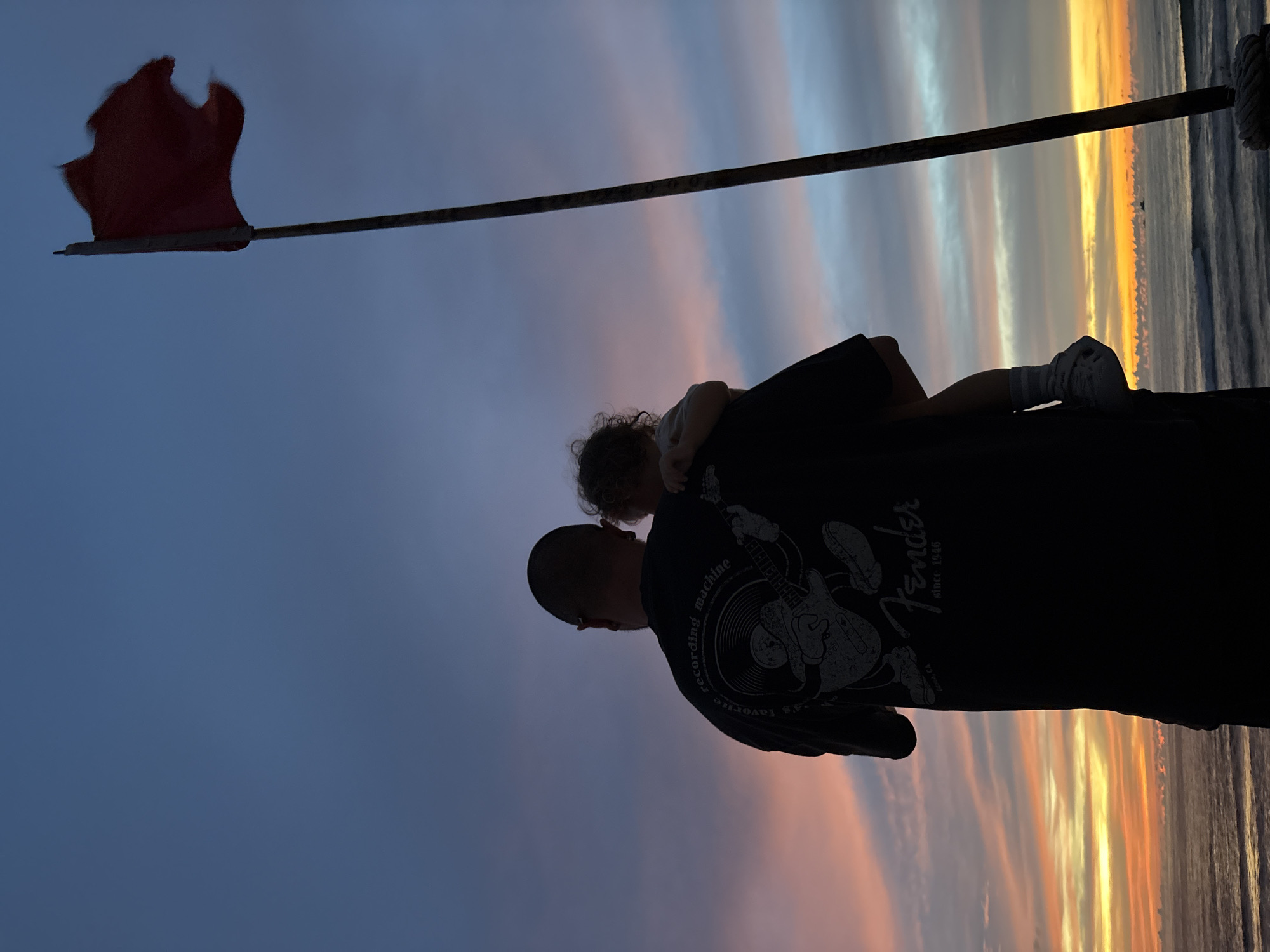 A father holds his child on his left side as they stand on a platform, silhouetted against a vibrant sunset over the ocean. A red flag flies from a pole to their right. The father is wearing a black t-shirt with a graphic print and the child has curly hair. The sky displays a gradient of colors, from deep blue at the top to orange and yellow at the horizon. The ocean exhibits gentle waves. The scene appears to be located on a beach in Choeng Thale, Thailand.