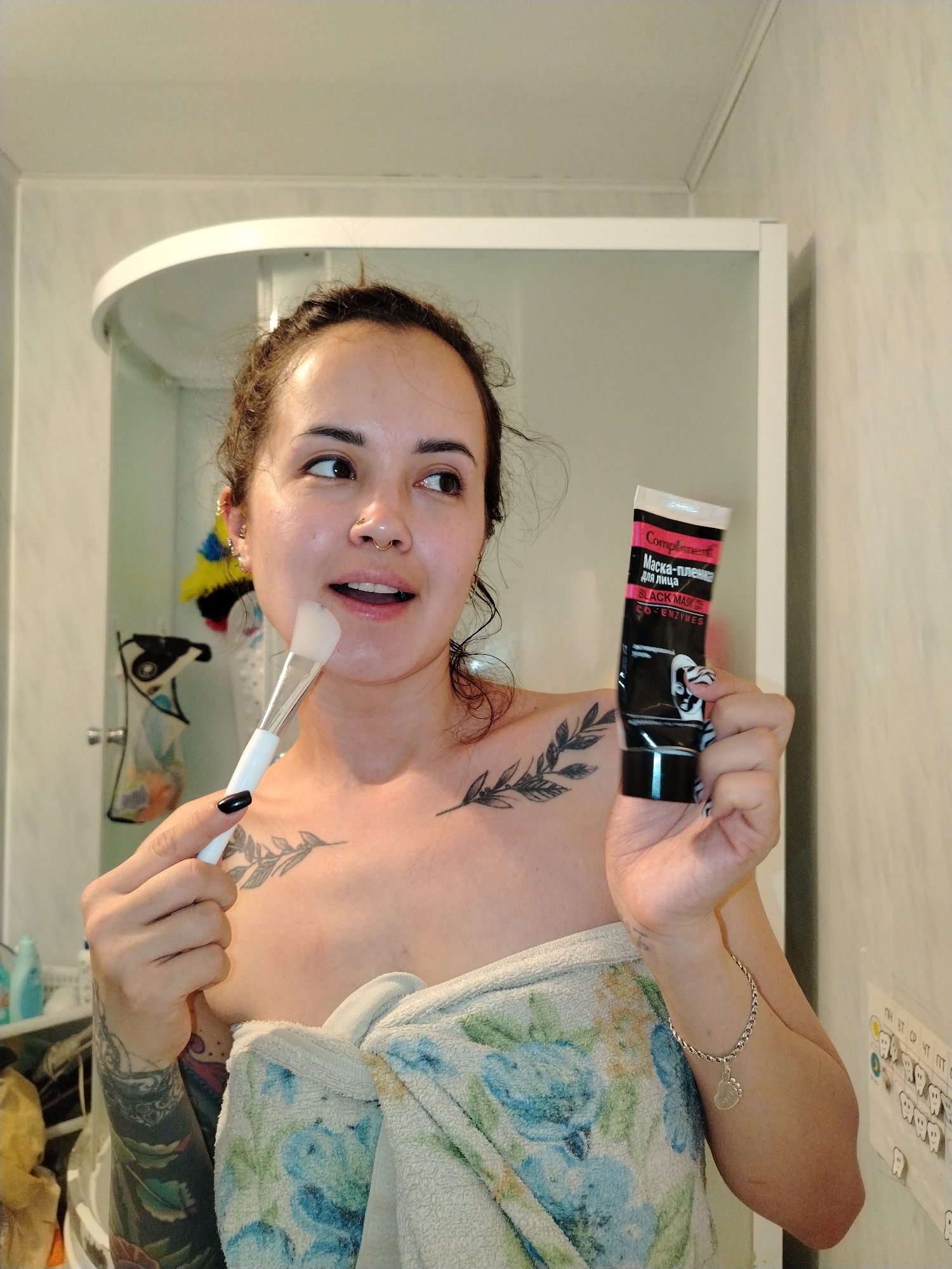 A woman with dark hair partially pulled back is in a bathroom. She is holding a white-handled makeup brush in her left hand and a black tube of "Complement Black Mask" in her right hand. She is partially wrapped in a floral-patterned towel. Tattoos are visible on her chest and left arm. A shower is behind her, and bathroom supplies are to her left. To the right is a wall with what appears to be a tooth chart. The location is Noviy, Russia.
