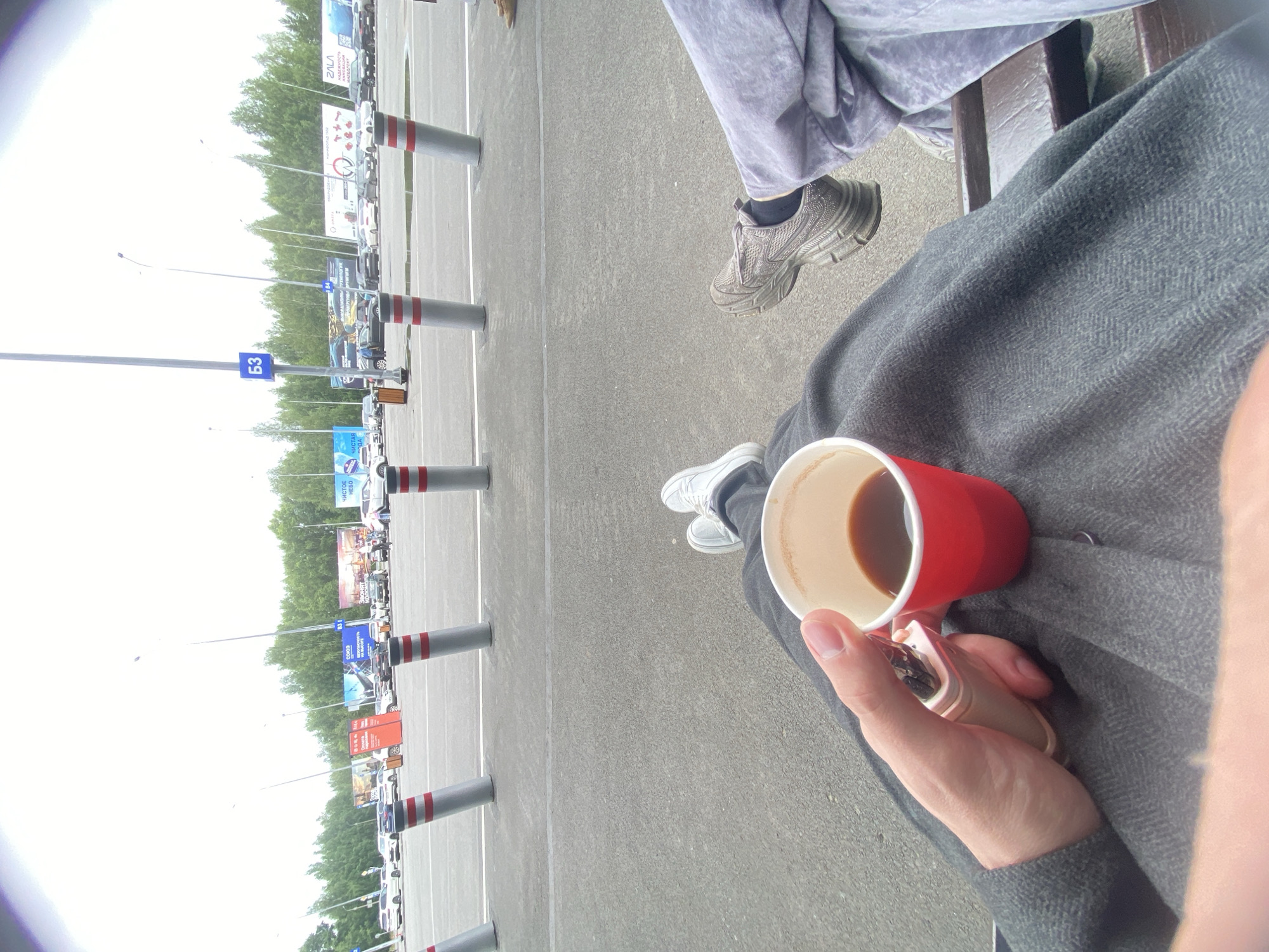 A medium shot shows a person holding a red paper cup of coffee in their left hand. They are seated on a bench, with their legs crossed. The person is wearing dark grey pants and a grey jacket. Their right leg is extended forward, and their right foot, clad in a grey sneaker, is visible. To the right, another person is also seated on the bench, wearing light purple pants and a grey sneaker. The background features a parking lot with several cars and large billboards. The scene is set outdoors, under an overcast sky, with trees visible in the distance. The location is identified as Novoye Martyanovo, Russia.