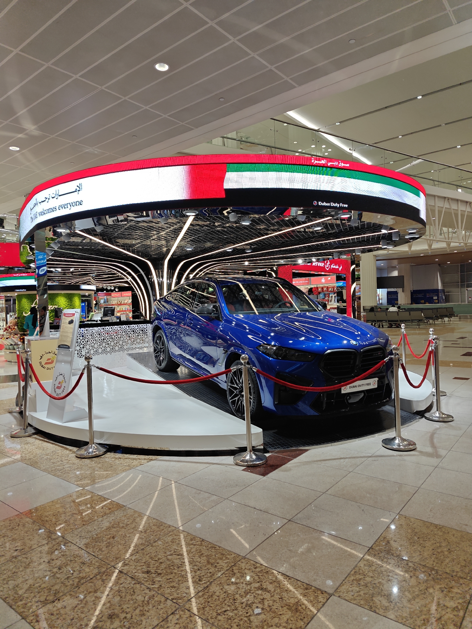 The image captures an indoor display featuring a vibrant blue BMW sedan parked on a raised white platform. The display is set within a modern, well-lit space that appears to be an airport terminal in Dubai, United Arab Emirates. Above the car, a large, curved LED screen displays the UAE flag colors and text in both Arabic and English, with "UAE welcomes everyone" clearly visible. The screen also shows "Dubai Duty Free," indicating the sponsor of the display. The area around the car is cordoned off with red velvet ropes and silver stanchions, suggesting it's an exhibit. In the background, there are glimpses of shops, seating areas with people, and architectural elements typical of an international airport concourse, including tiled flooring and suspended ceilings with lighting. The overall scene suggests a promotional event or exhibition within a busy travel hub.
