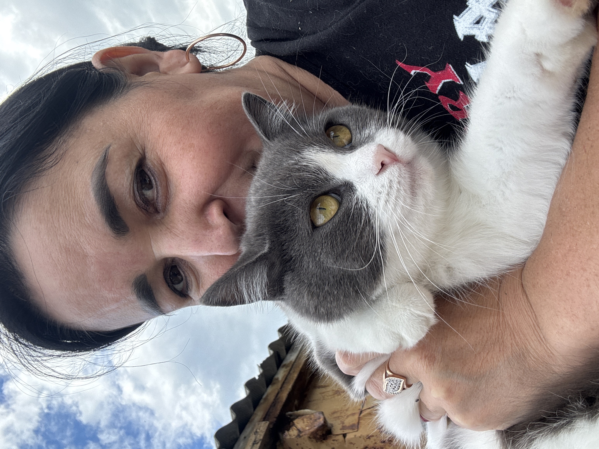 A woman with dark hair and hoop earrings is holding a gray and white cat in Starokhalilovo, Russia. The woman is wearing a black t-shirt. She is wearing a ring on her left ring finger. The cat has yellow eyes and a pink nose. In the background, there is a wooden structure with a corrugated metal roof, and a cloudy sky.