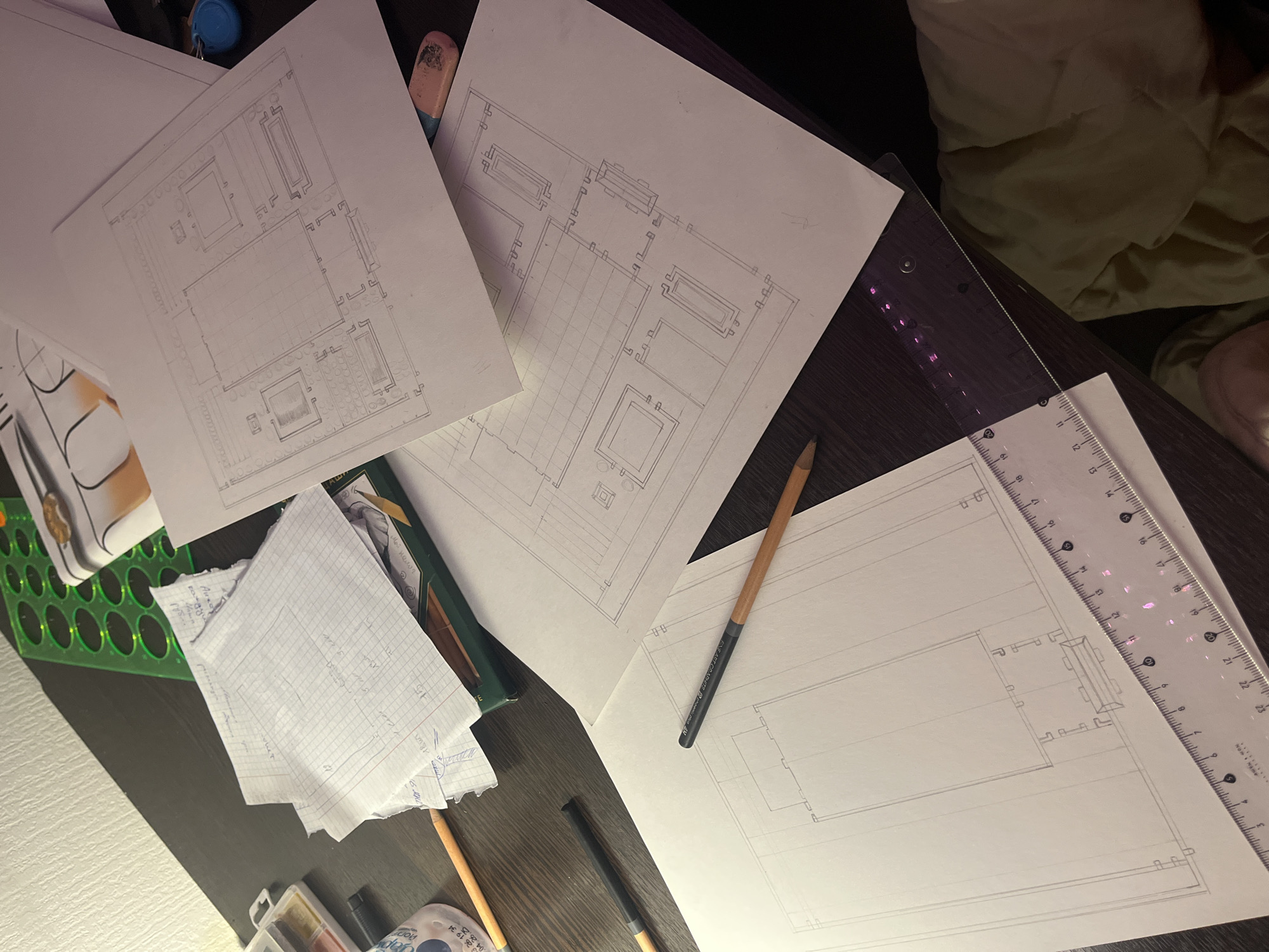 This image shows a desk with architectural drawings, pencils, a ruler, and other drafting tools. The drawings appear to be floor plans or elevations of buildings, with detailed layouts and dimensions indicated. The desk is dark, and there is a bed or cushion visible in the background. The scene is cluttered, with various items scattered around the desk, suggesting an active work environment. There's nothing in the image to indicate it's in Saint Petersburg, Russia.